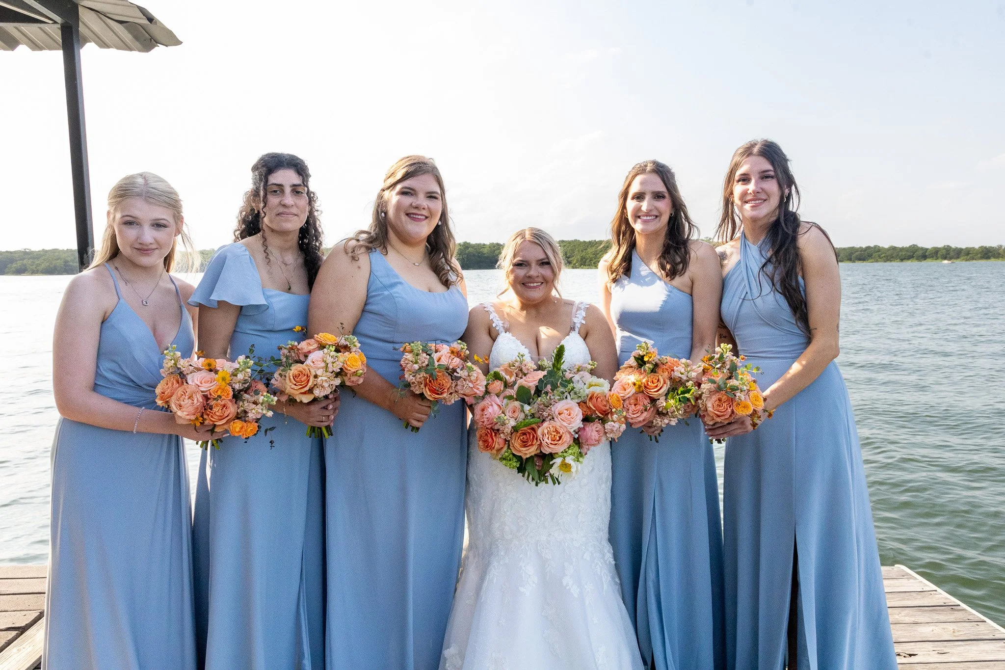 Bride in white wedding dress with six bridesmaids in light blue dresses, standing on a dock by a body of water, holding pink and peach bouquets, smiling at the camera.
