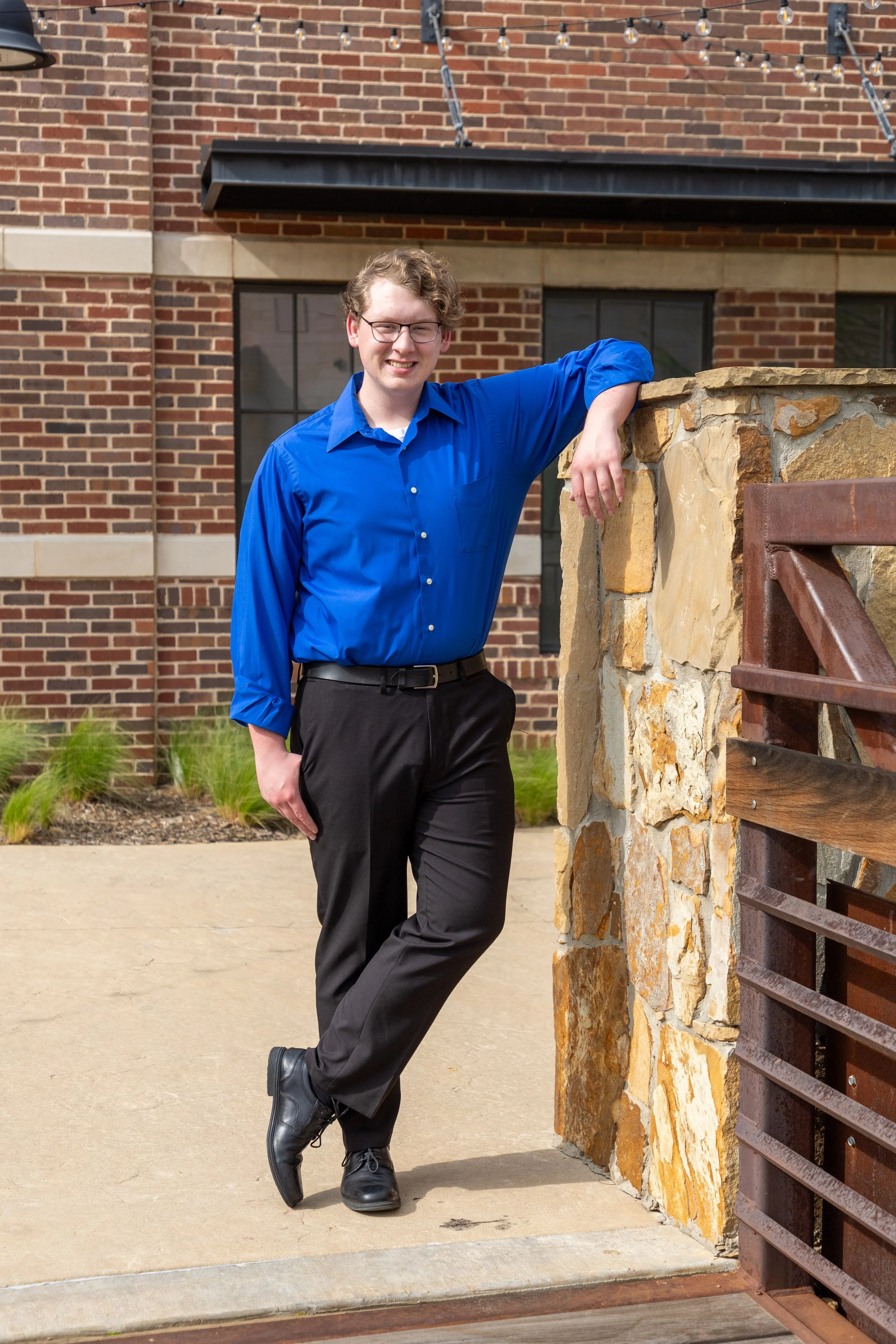 Young man with glasses in a blue shirt and black pants leaning against a stone wall outside a brick building, smiling at the camera.