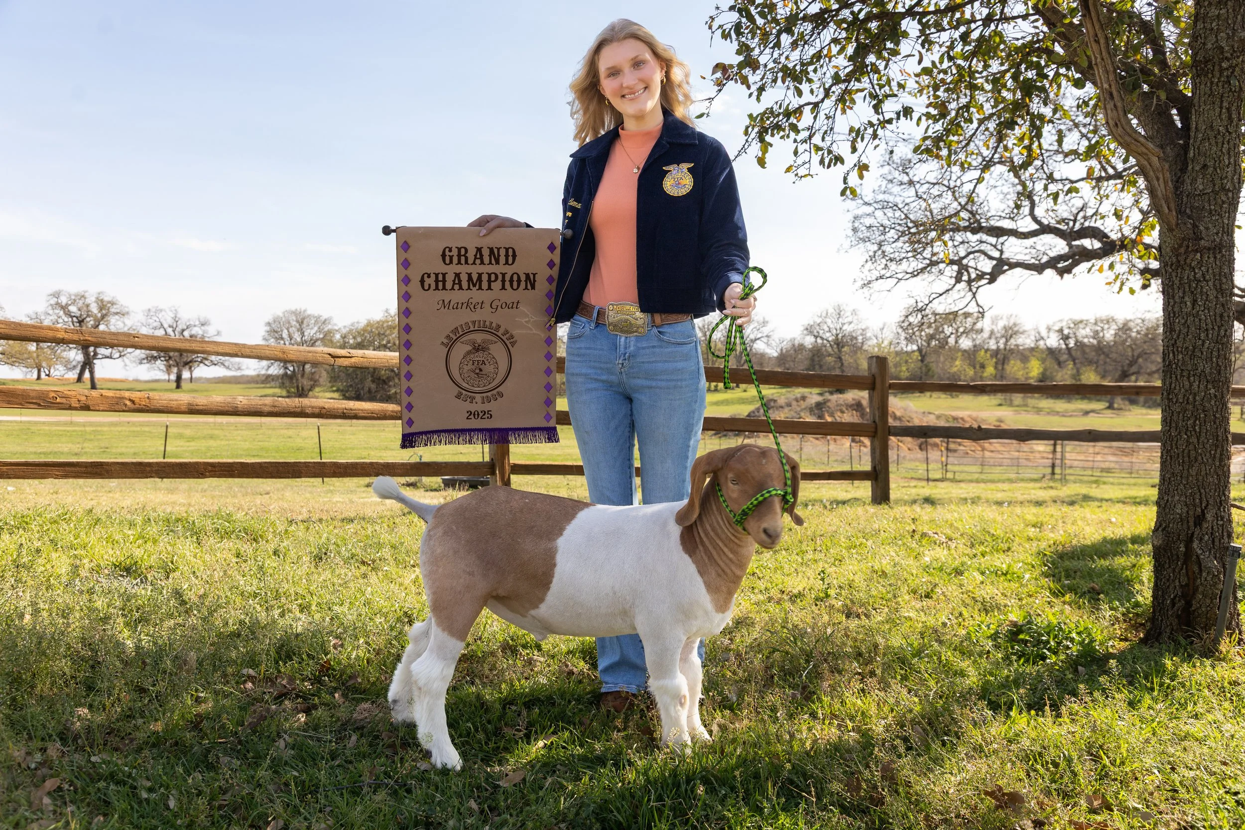 A young woman standing outdoors in a grassy field, holding a small goat on a green leash. She is smiling and wearing a dark blue FFA jacket, a pink shirt, and blue jeans. She holds a purple and white banner that reads "Grand Champion Market Goat" wit
