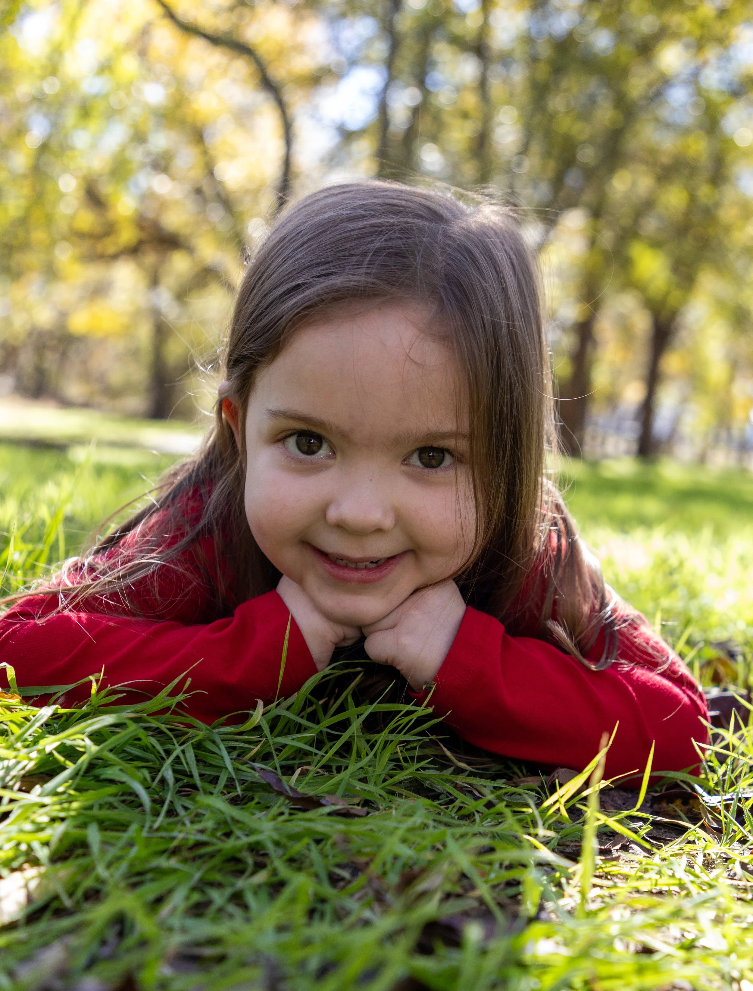 Young girl lying on grass in a park during fall, wearing a red shirt, with trees and yellow leaves in the background.