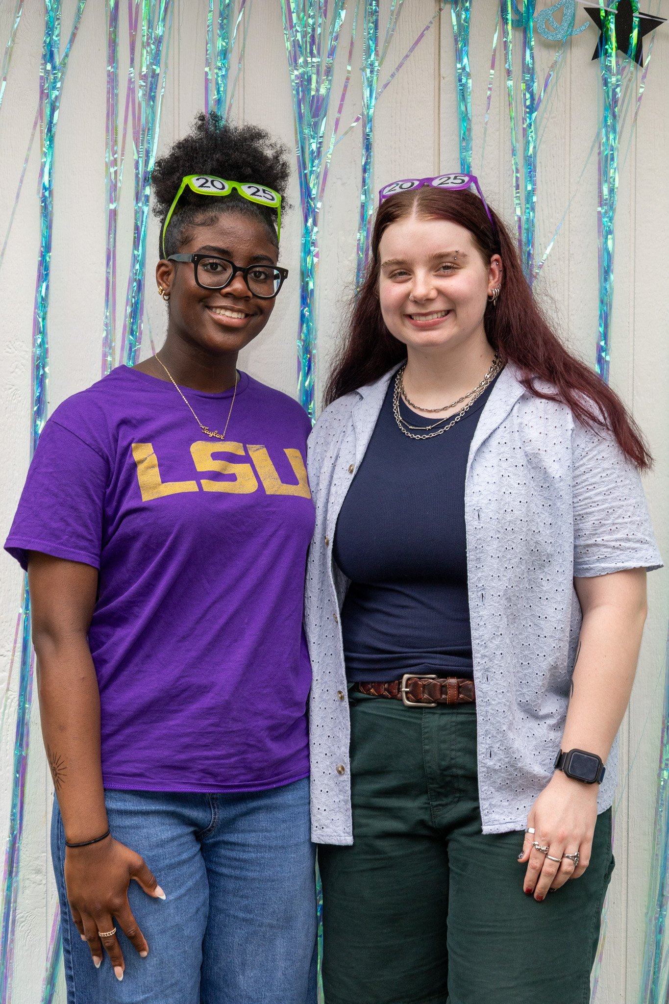 Two young women standing together at a graduation celebration, smiling, with colorful metallic streamers hanging behind them. One is wearing a purple LSU t-shirt, glasses, and multicolored sunglasses on her head. The other is wearing a navy blue shir
