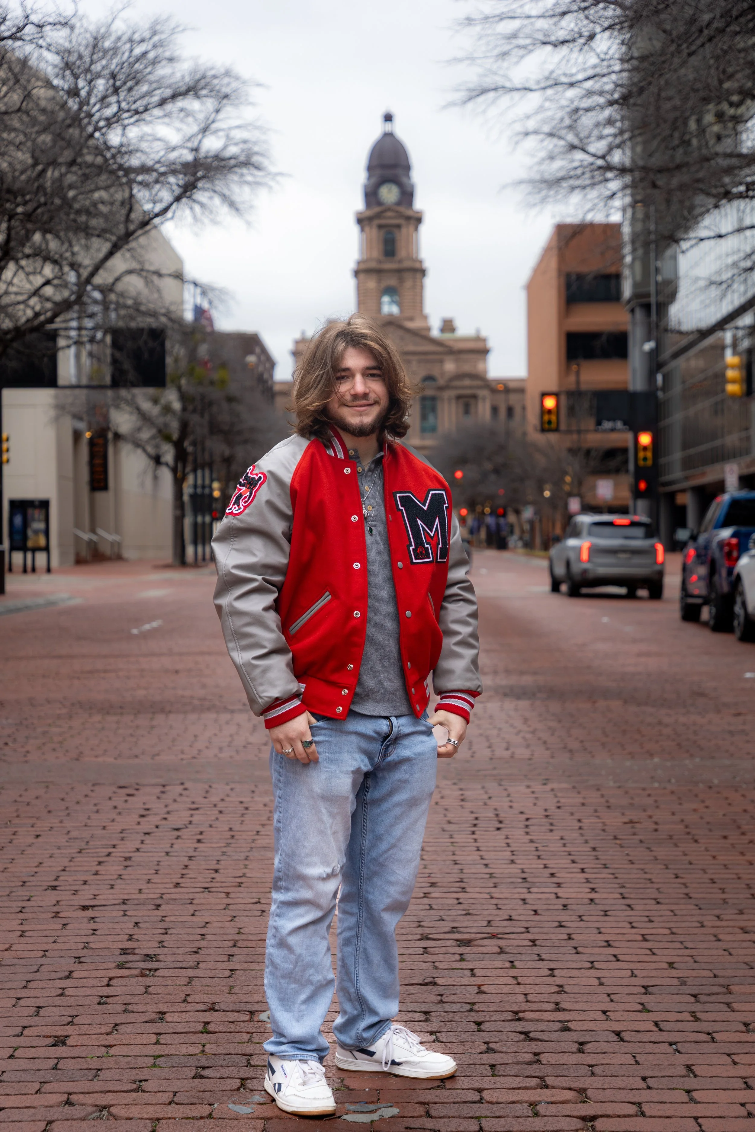 A young man with shoulder-length hair standing on a brick-paved street, wearing a red and gray varsity jacket, light blue jeans, and white sneakers. There are cars and buildings with leafless trees in the background.