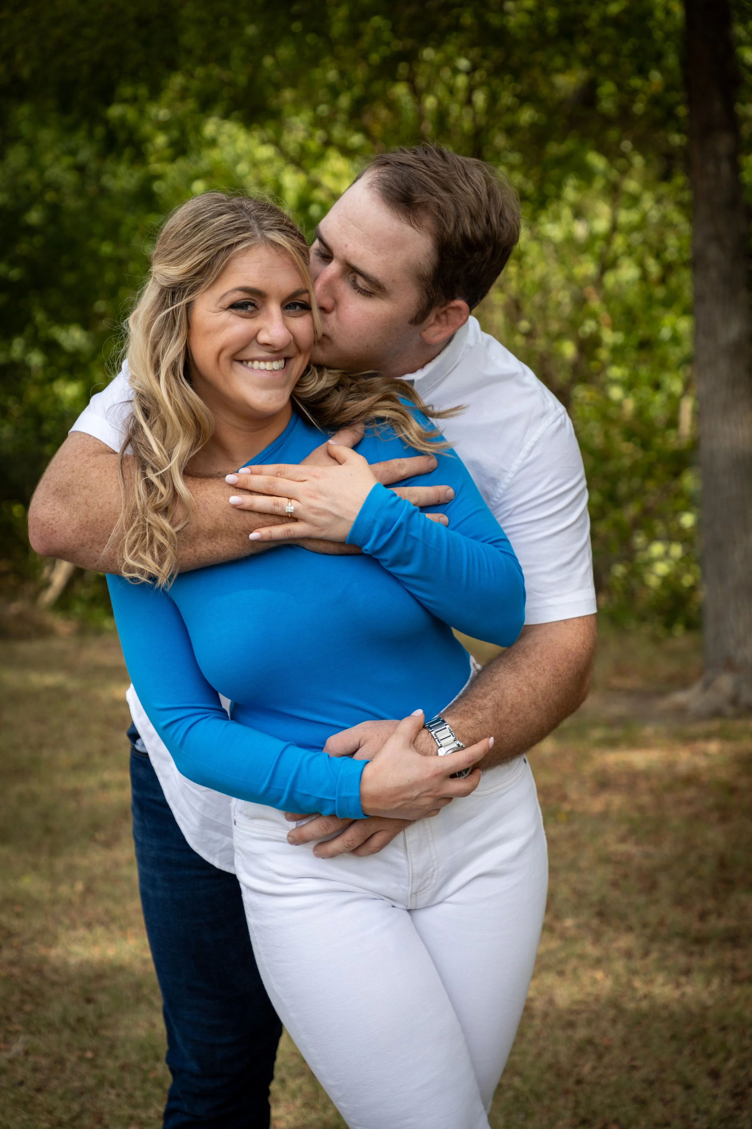 A man giving a woman a hug from behind outdoors, with trees and green foliage in the background. The woman is smiling at the camera, wearing a blue long-sleeve shirt and white pants. The man, in a white shirt, is kissing her cheek.