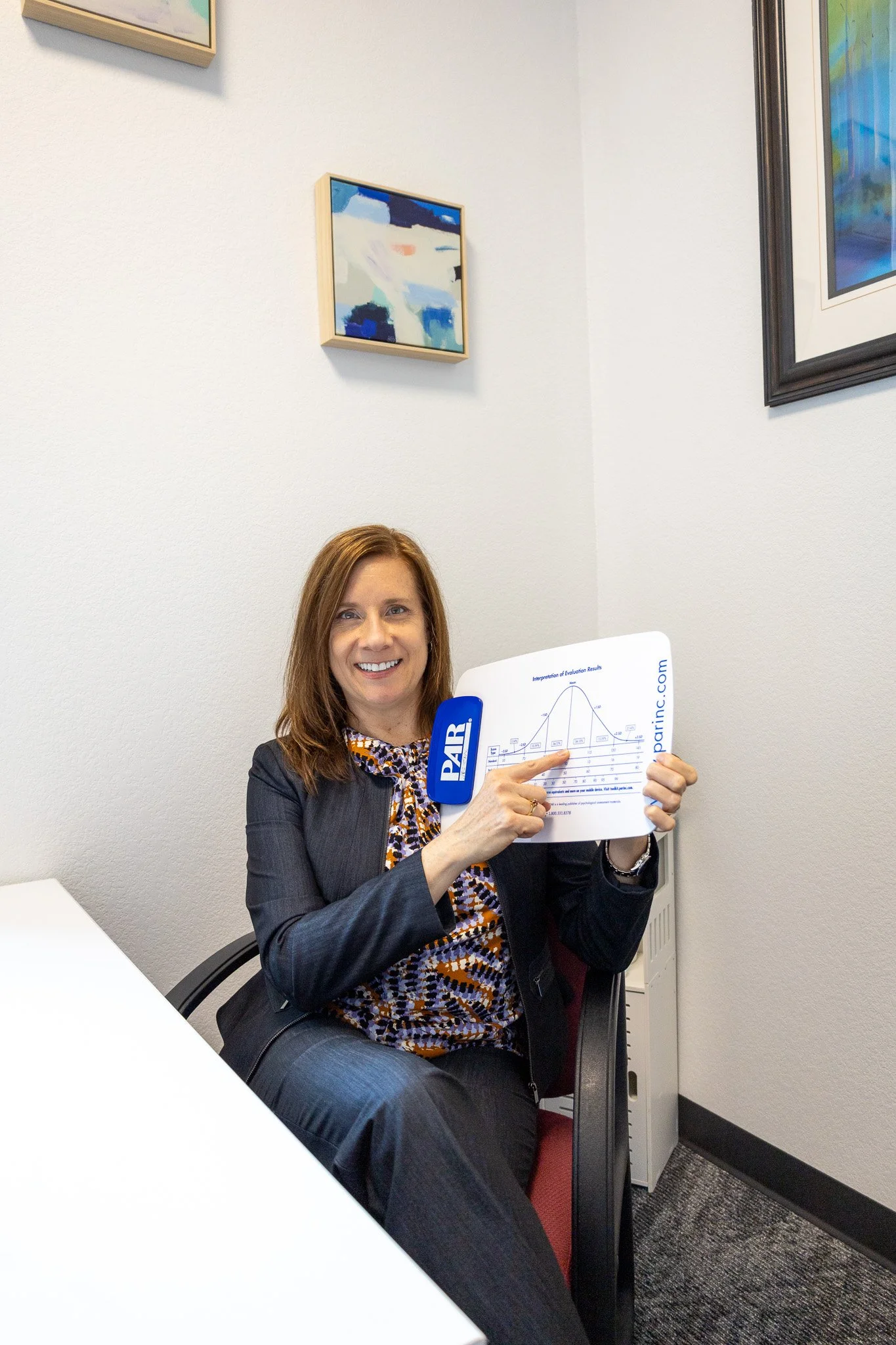 A woman seated in an office, holding a sheet with a chart, pointing at it, smiling, with framed abstract art on the wall behind her.