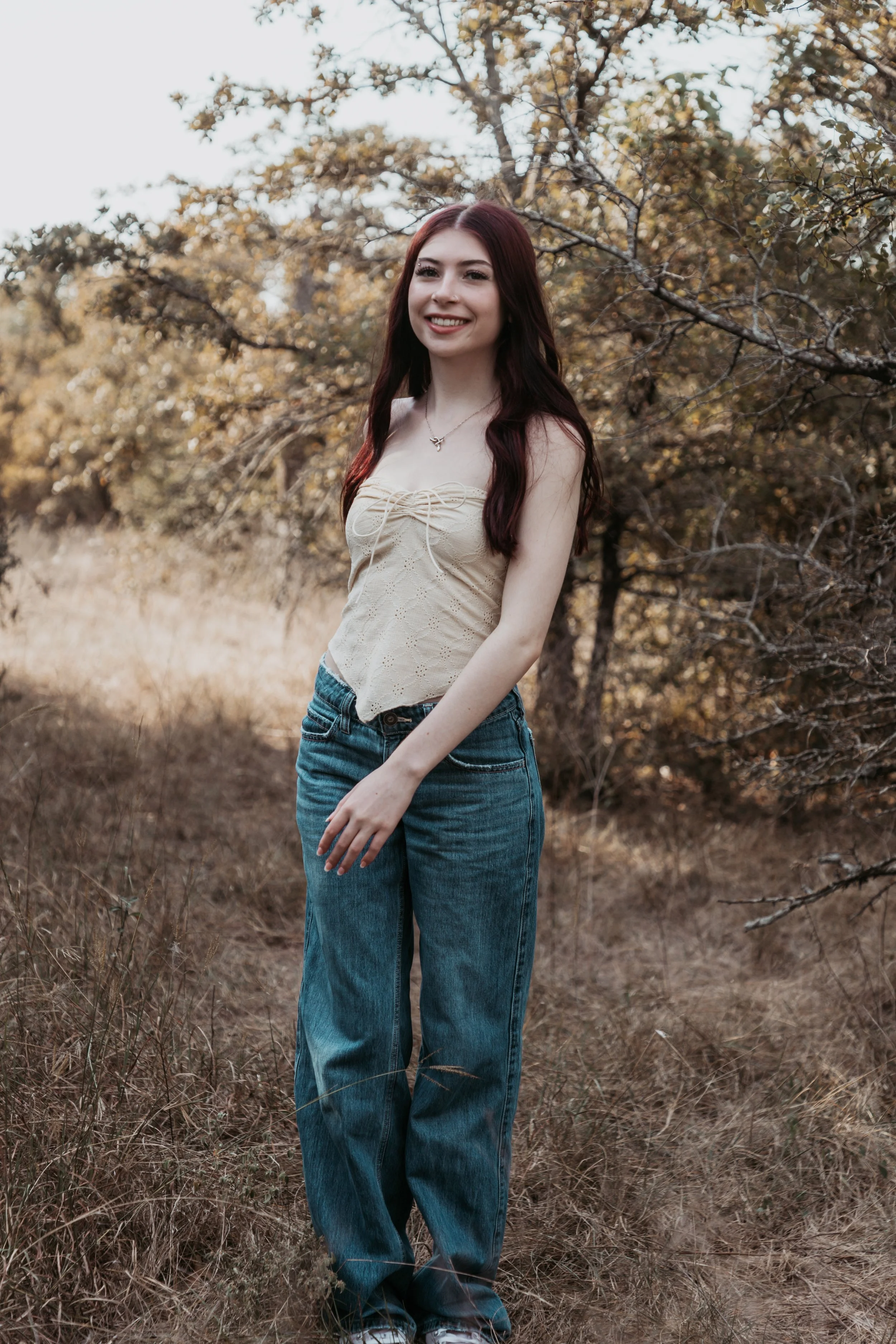 A young woman with long dark red hair and fair skin, wearing a beige strapless top and blue jeans, standing outdoors on a grassy field with trees in the background, smiling at the camera.