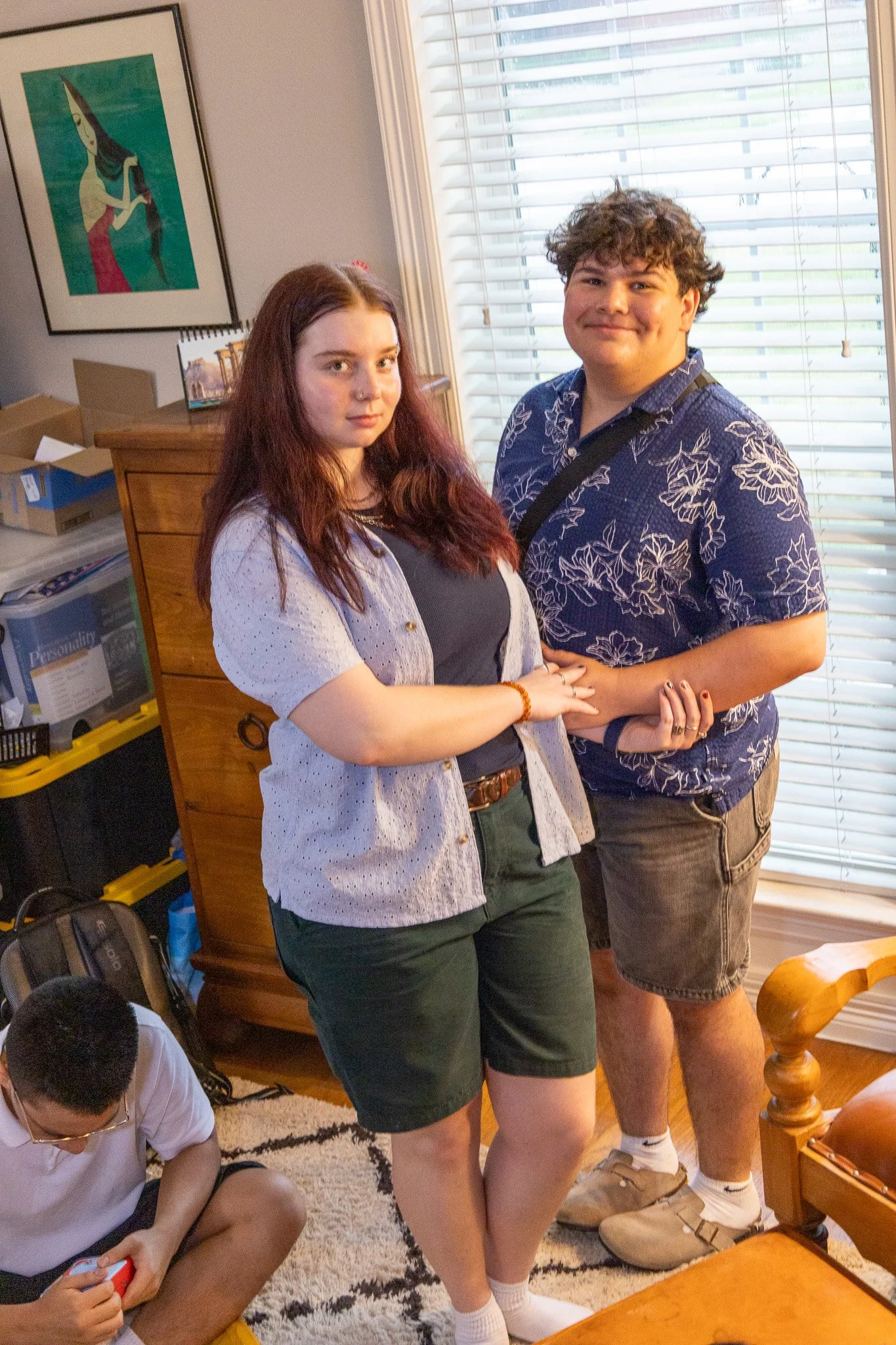 Two people stand close together indoors, holding hands, with a woman with long red hair and a man with curly hair smiling. A boy is sitting on the floor nearby looking at a device.