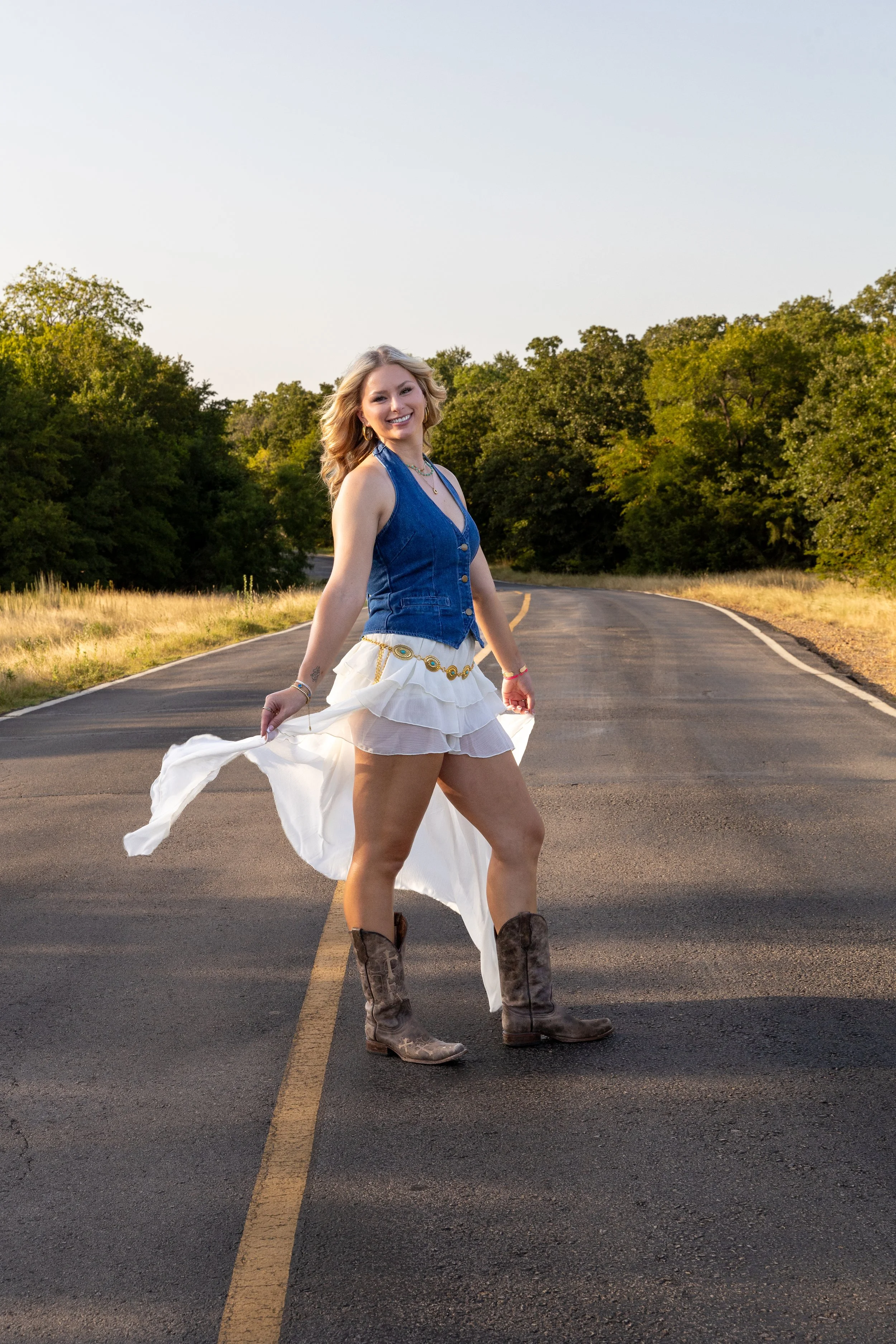 A woman wearing cowboy boots, a denim vest, and a white skirt stands in the middle of a deserted road with trees on both sides, smiling at the camera during sunset.