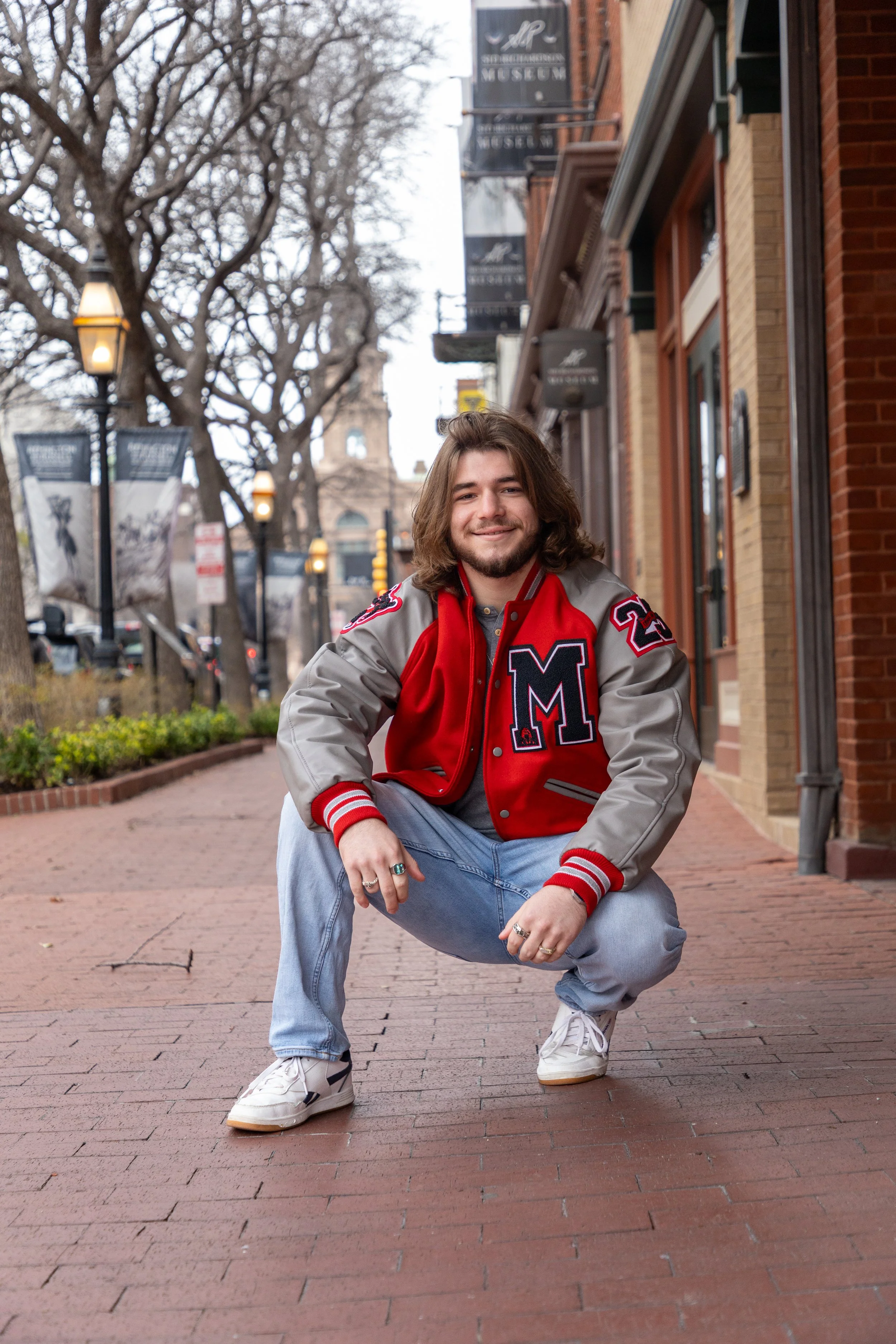 Young man crouching on a brick sidewalk in an urban area, wearing a red and gray varsity jacket, light blue jeans, and white sneakers, with trees and historic buildings in the background.