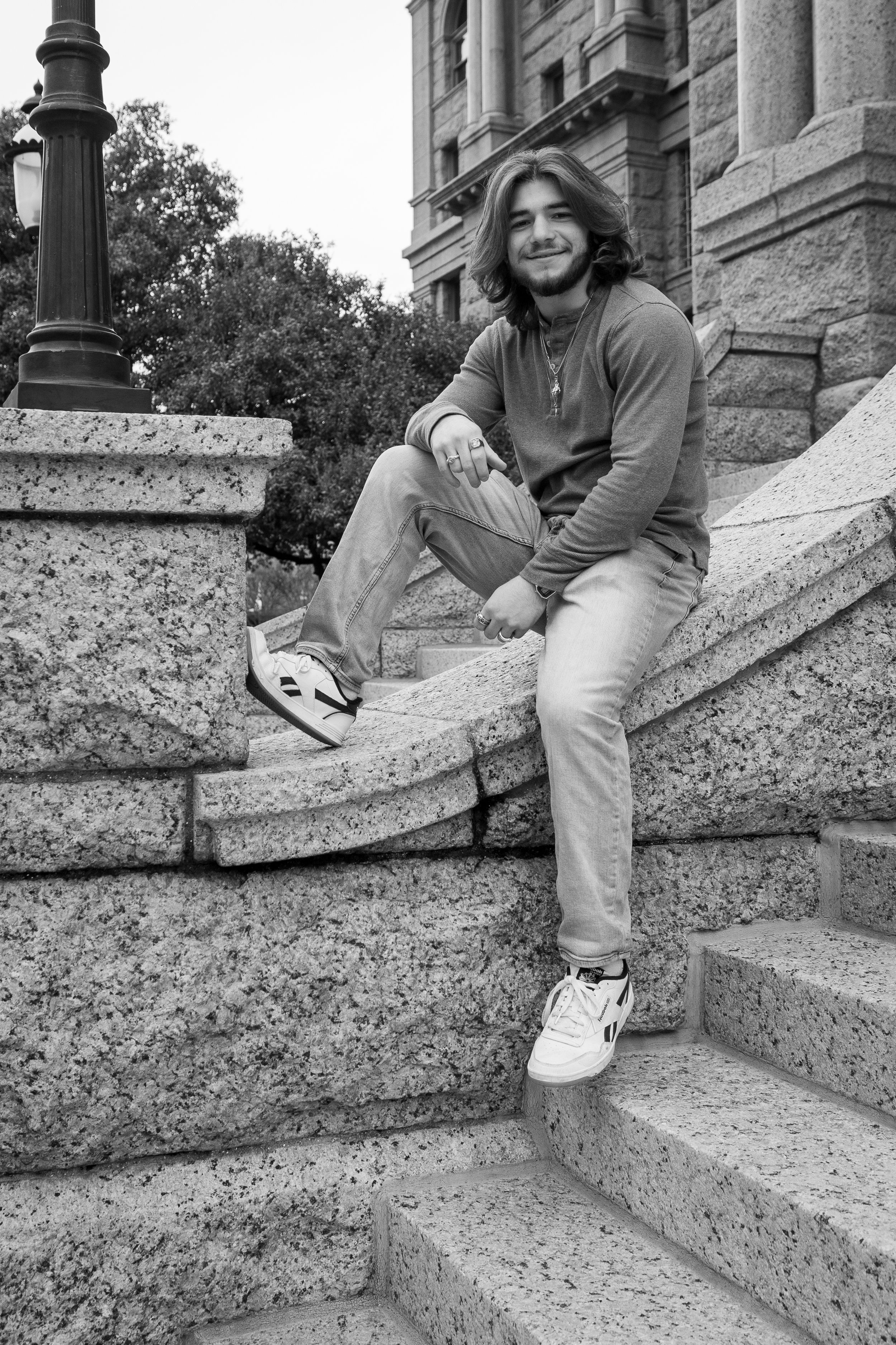 A young man with shoulder-length hair and a beard, sitting on stone steps outside a historic building, smiling at the camera.