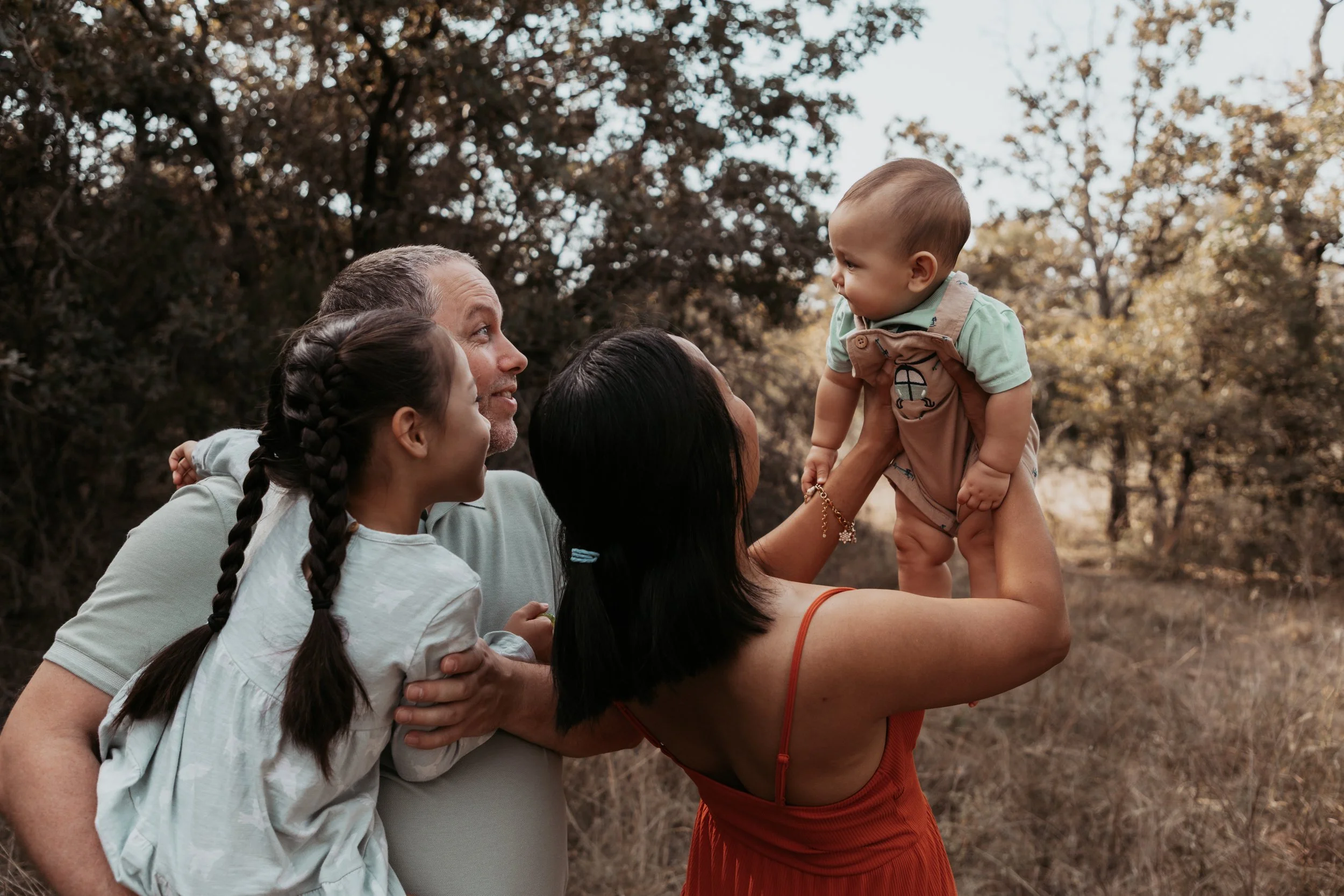 A multiracial family with two adults and two children outdoors in a wooded area, with one adult lifting a toddler in the air. They are enjoying a moment of togetherness.