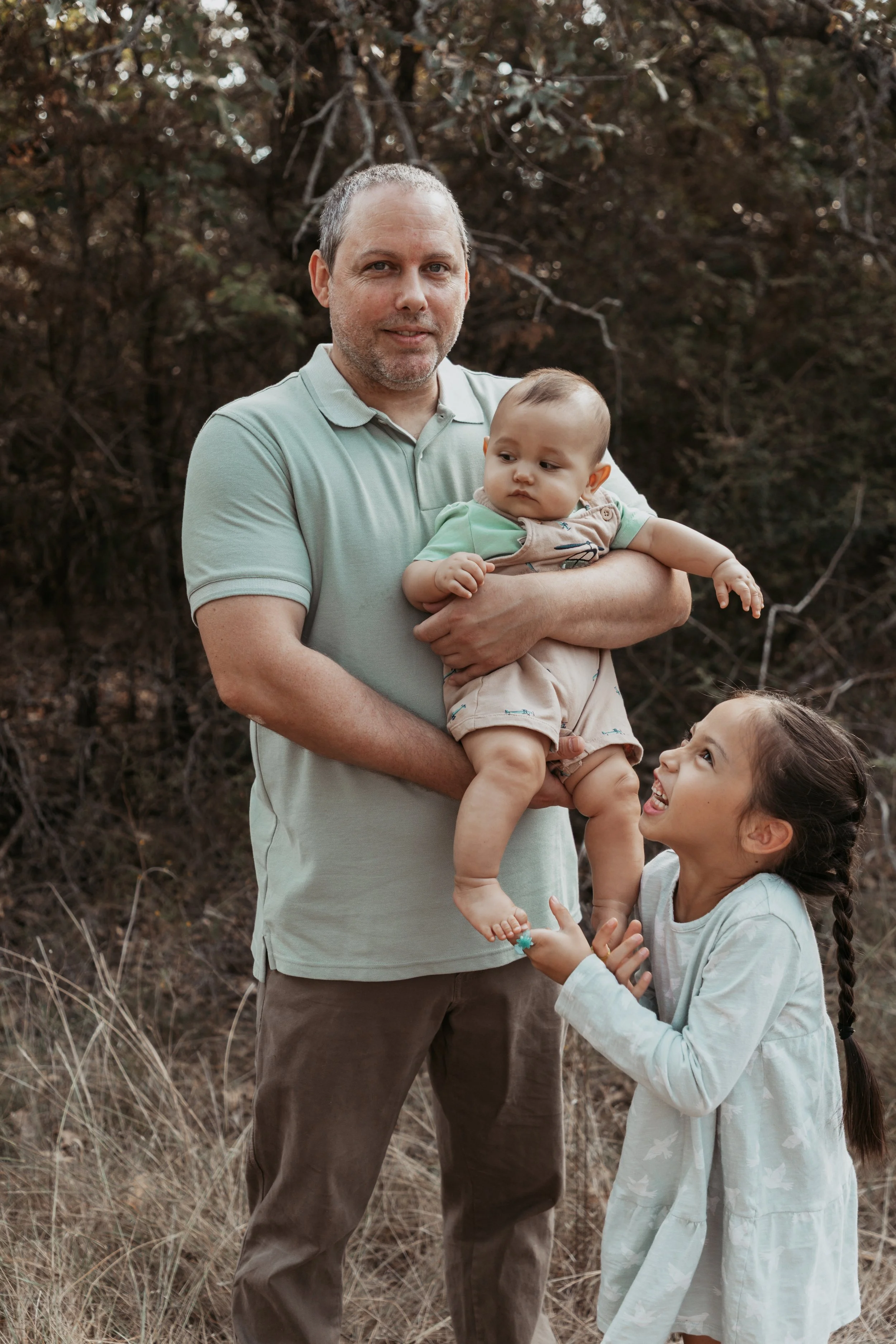 A man holding a baby boy in an outdoor setting with dry grass, while a young girl with a braid looks up at the baby and smiles.
