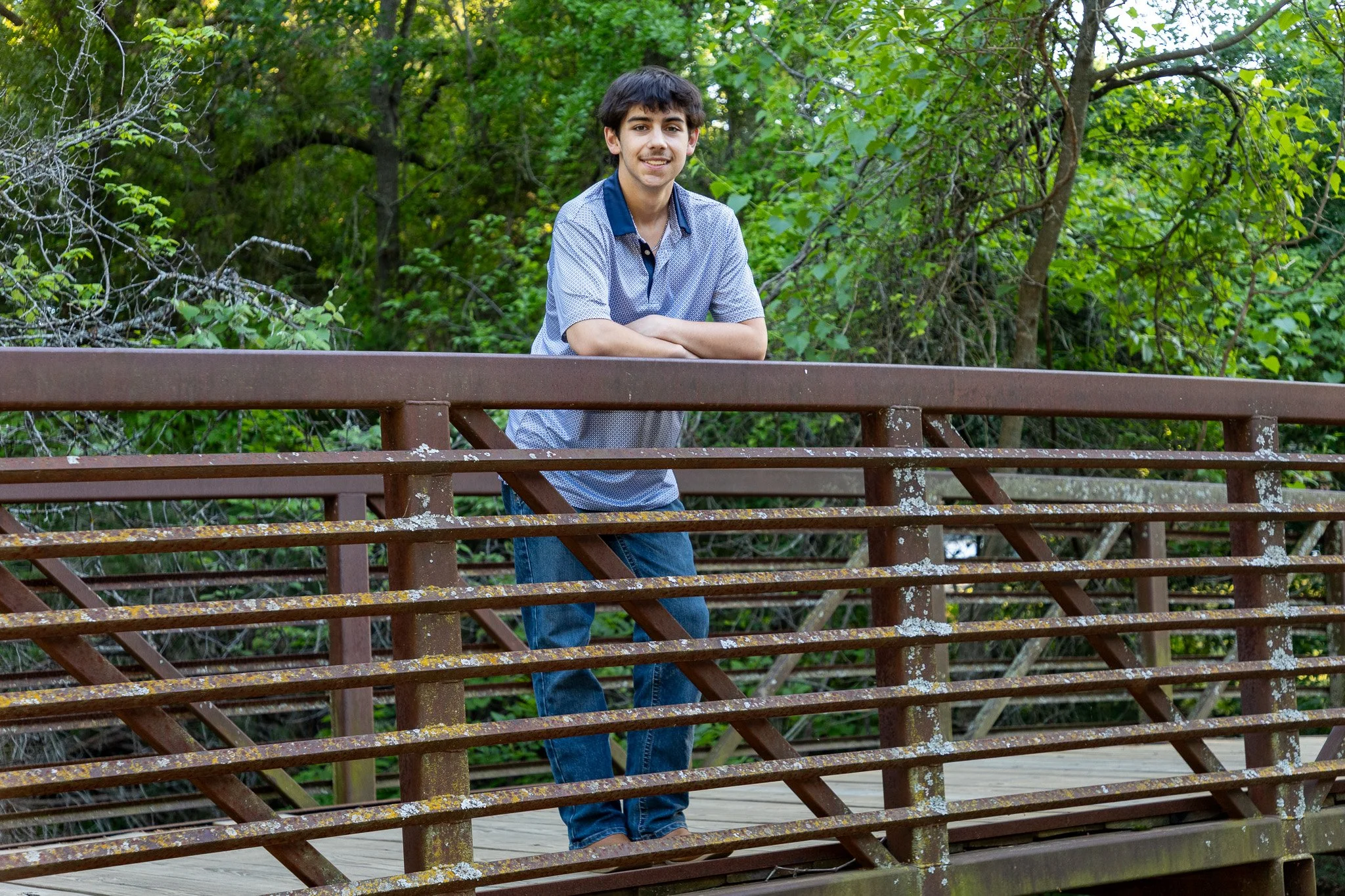 A young man with brown hair, wearing a light blue patterned shirt and blue jeans, is leaning on a brown metal railing on a wooden bridge, surrounded by green trees and foliage.