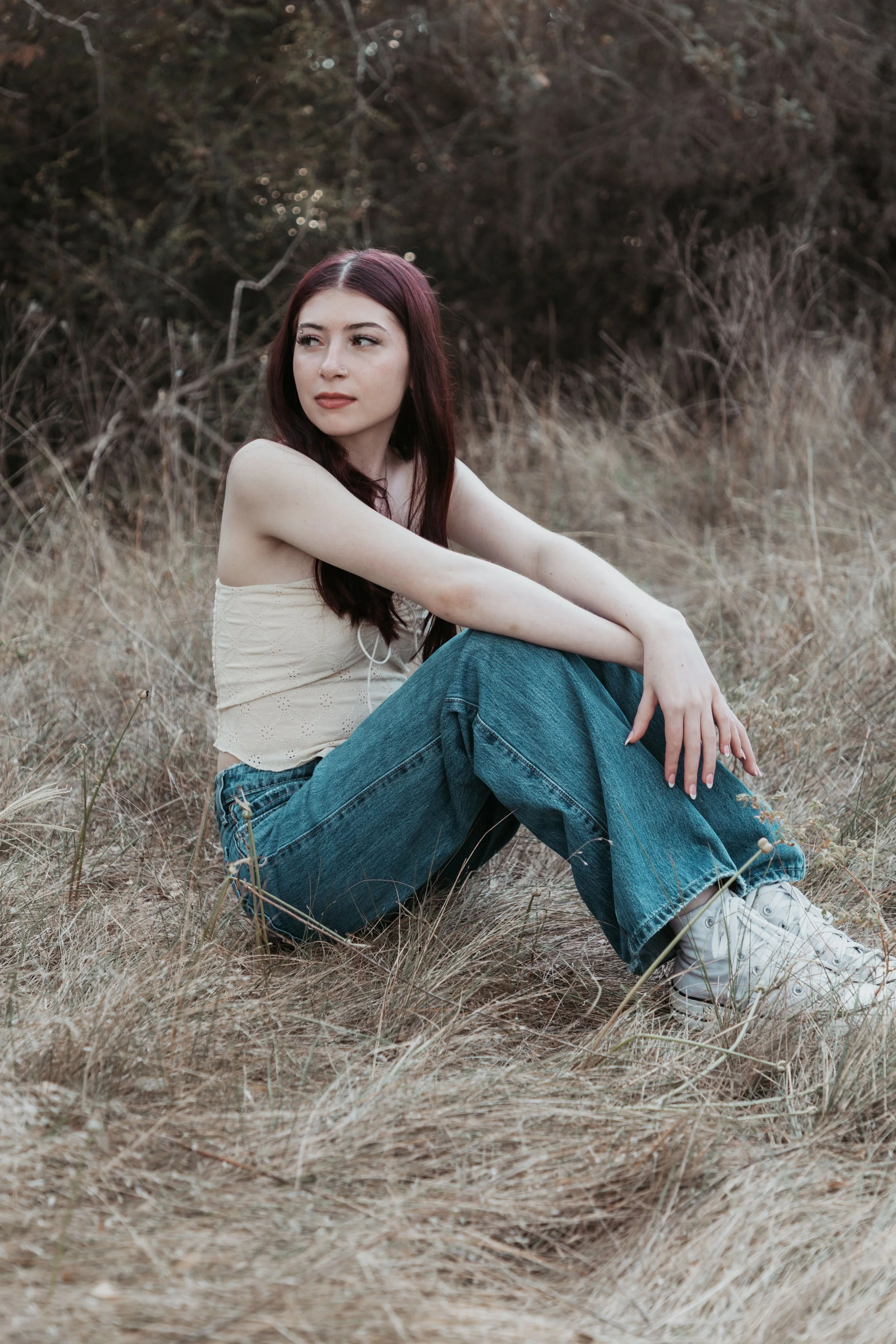 A young woman with long red hair sitting in dry grass in a natural outdoor setting, wearing a beige sleeveless top, blue jeans, and white sneakers, with a background of trees and branches.