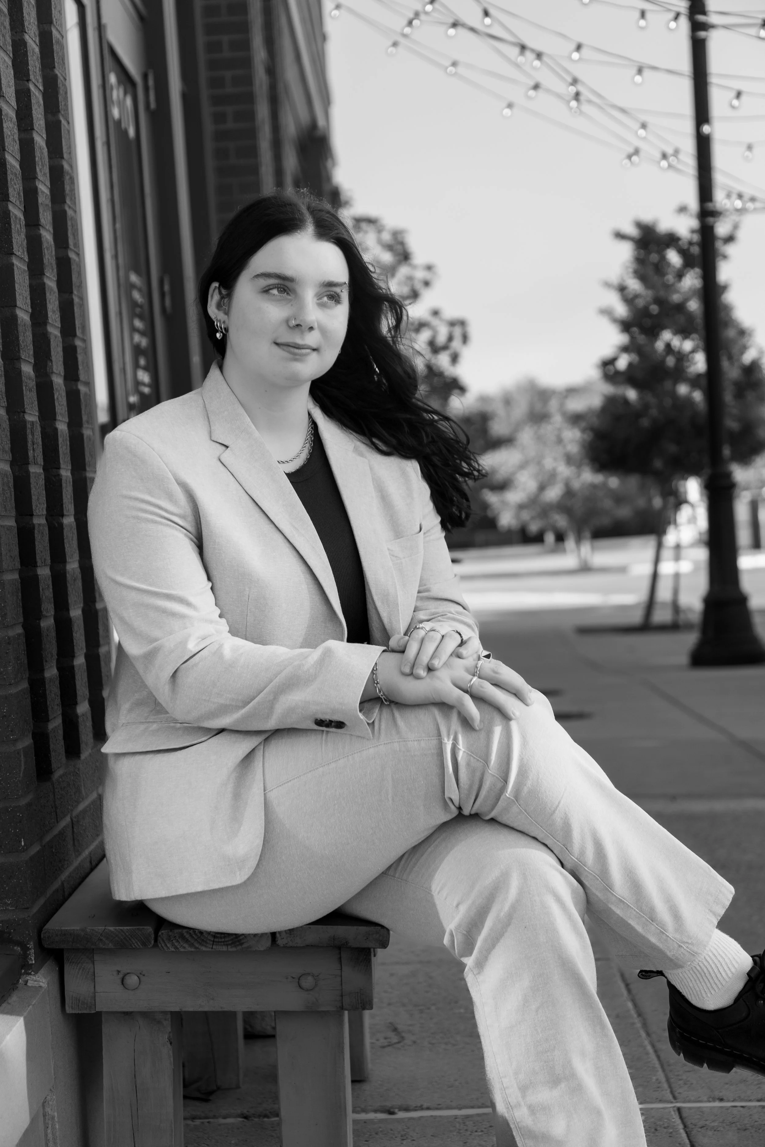 A young woman with long dark hair, dressed in a light-colored suit, sitting on a wooden bench outside on a city sidewalk. She is looking off to the side, with her hands resting on her lap. There are string lights hanging above and trees in the backgr