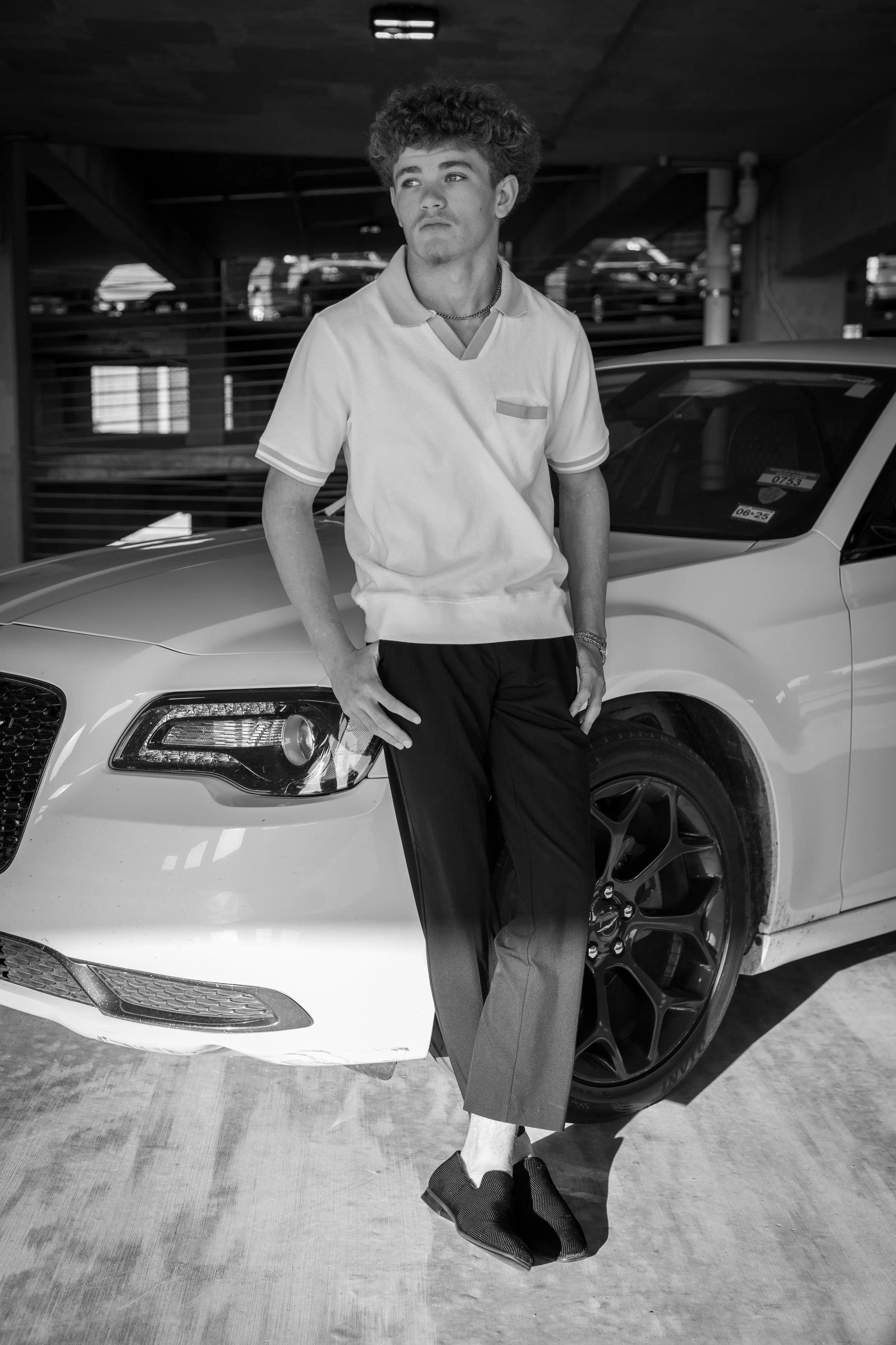 A young man with curly hair leaning against a white luxury car in an indoor parking garage.