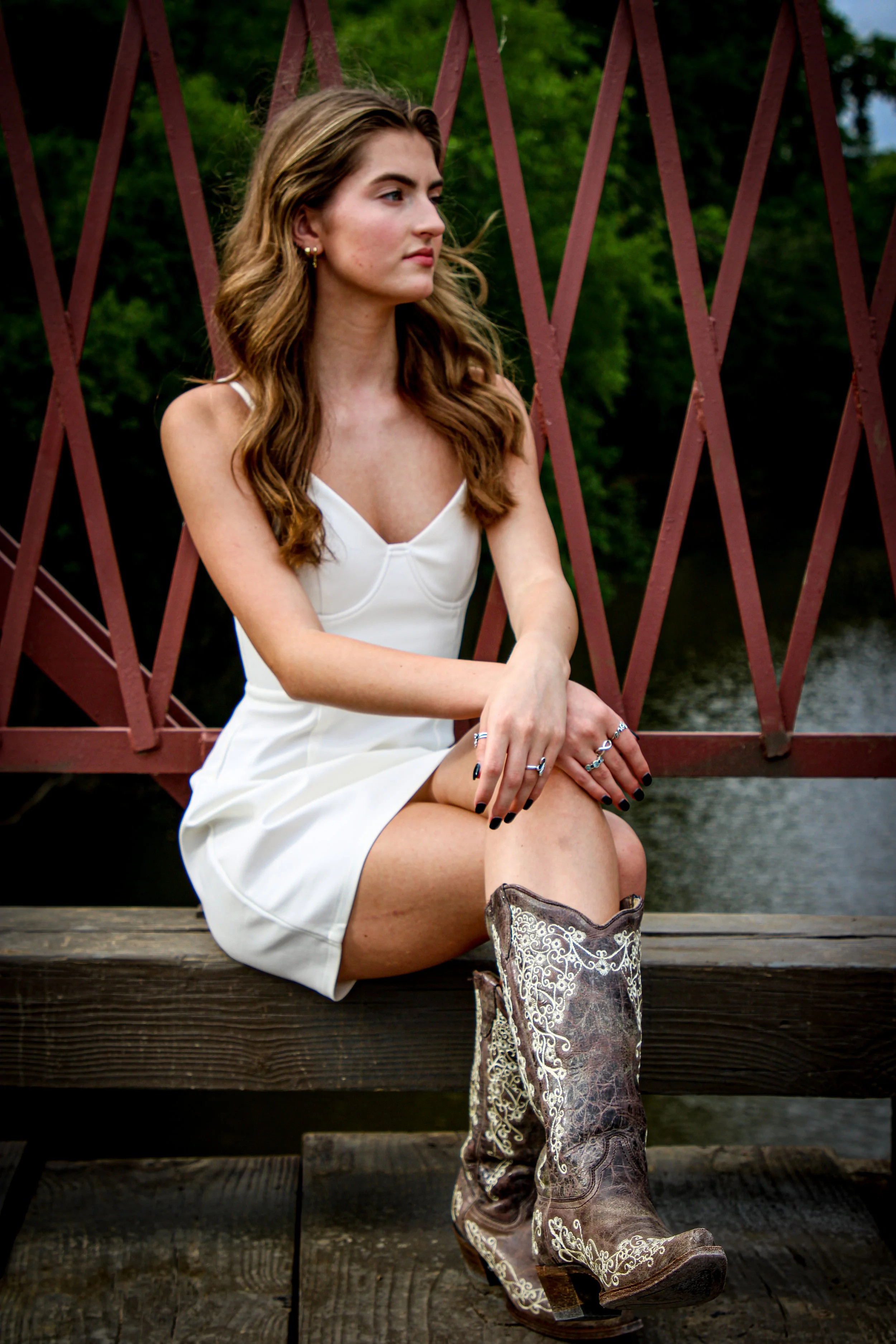 A young woman with long, wavy brown hair sitting on a wooden bridge, wearing a white dress and cowboy boots with embroidered details, with trees and water in the background.