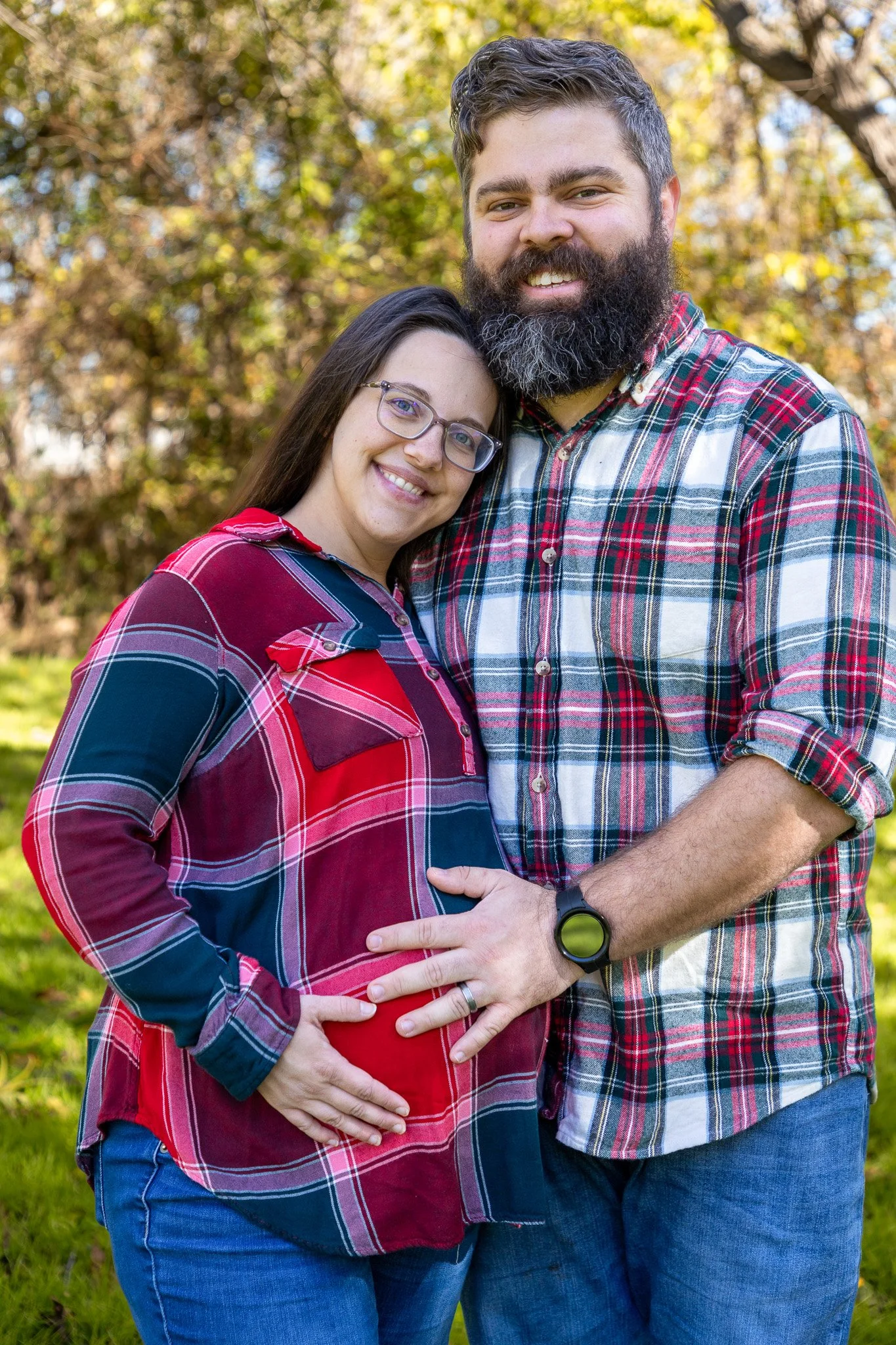 A smiling pregnant woman wearing glasses and a red plaid shirt stands next to a bearded man in a plaid shirt. They are embracing outdoors in a park with autumn leaves in the background.