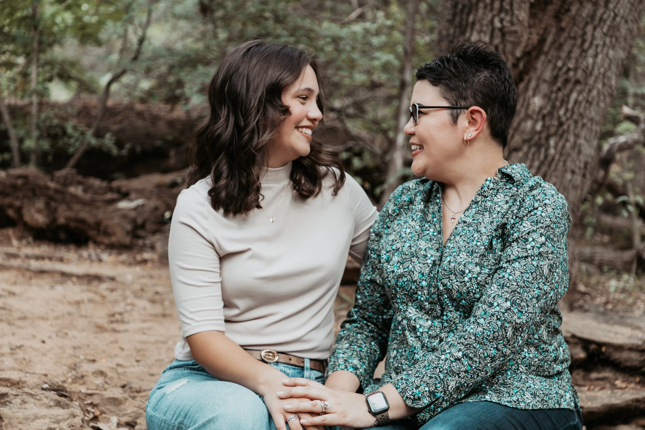 Two women sitting outdoors in a wooded area, smiling and holding hands, looking at each other affectionately.