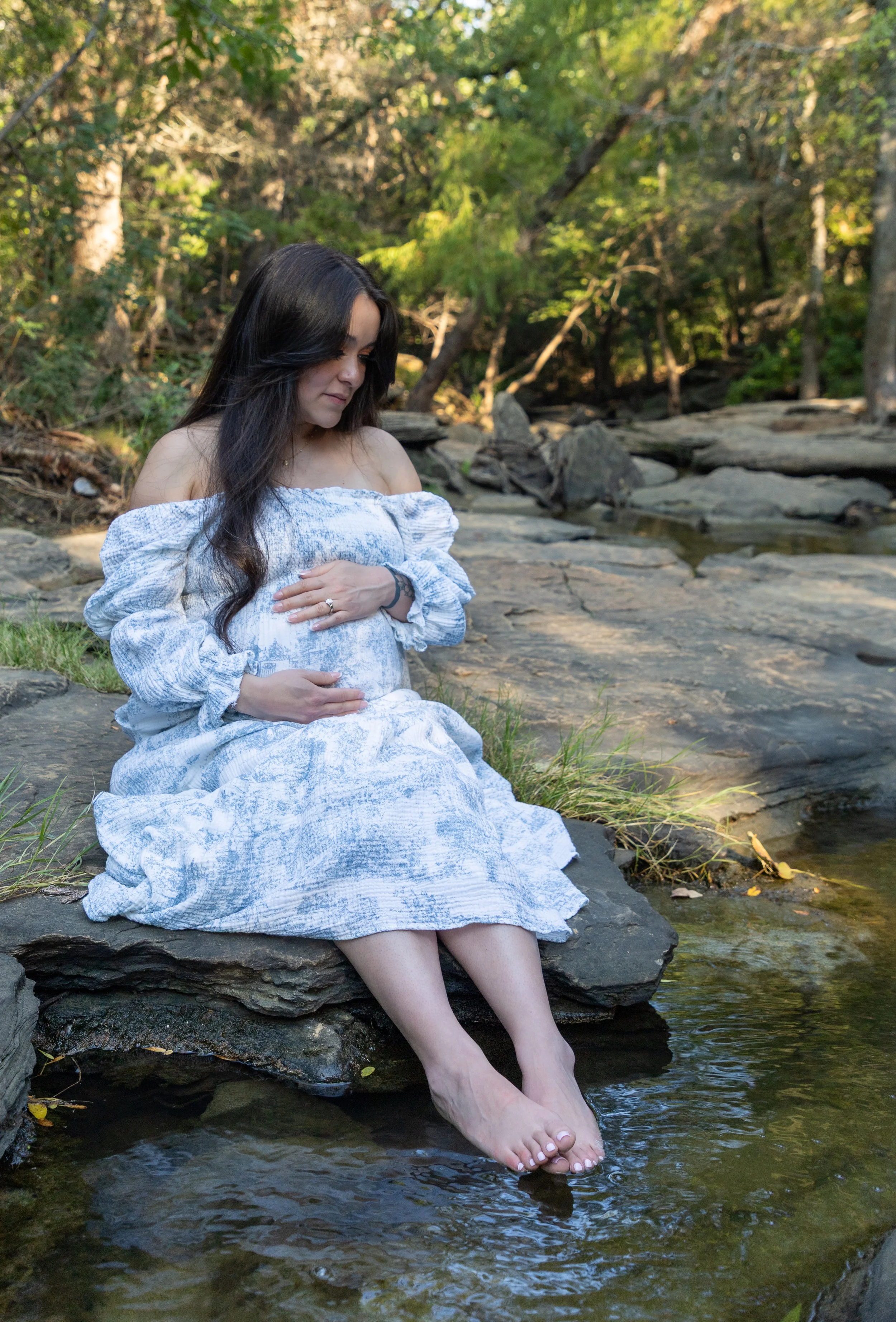 A woman in a white dress sitting on a rock by a stream, holding her pregnant belly with both hands, in a forest setting.