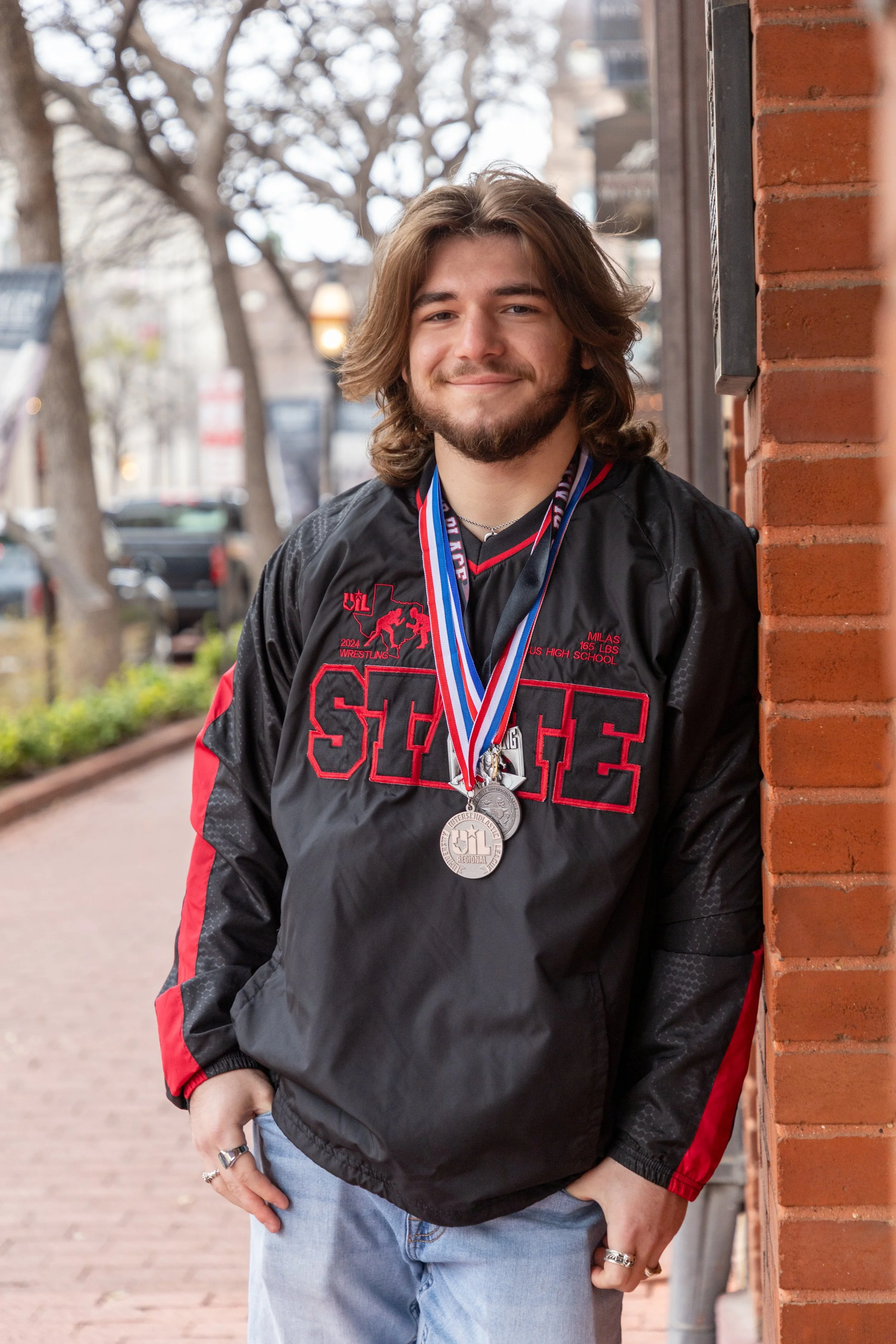 A young man with long hair, a beard, and a smile, wearing a black and red sports jacket with "Spartans" embroidered on it, standing outdoors near a brick wall, with medals around his neck.