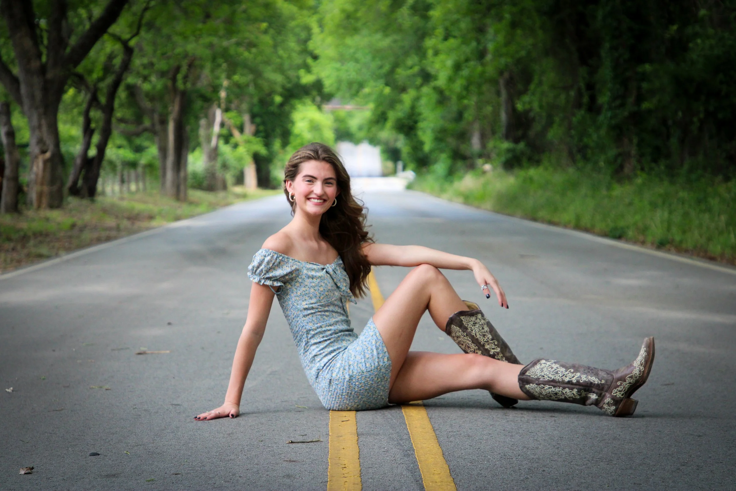 A young woman with long brown hair, wearing a blue patterned dress and cowboy boots, sitting on a deserted country road with green trees on either side, smiling at the camera.