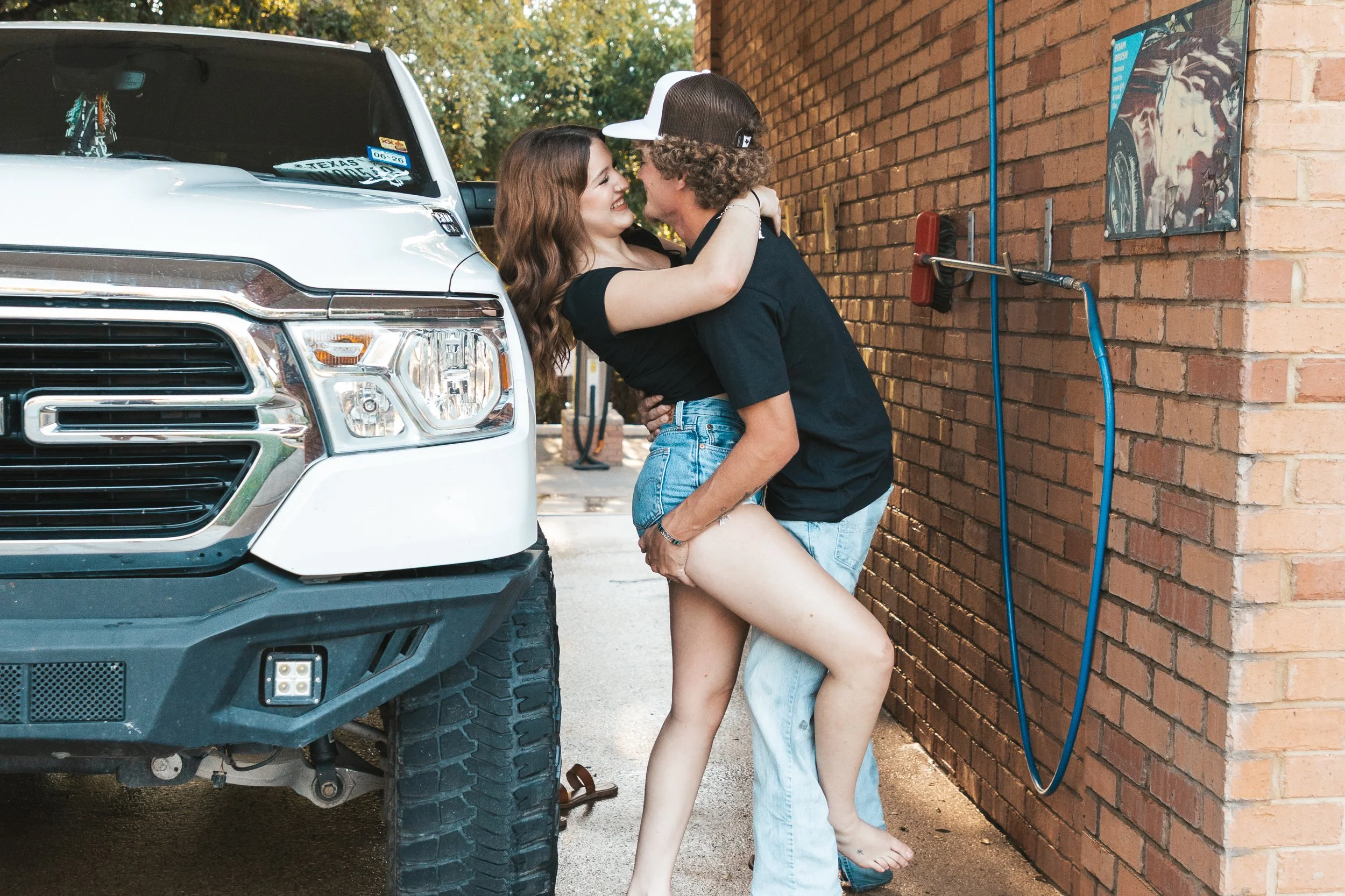 A young couple embracing and smiling outside near a brick wall with a white truck nearby.