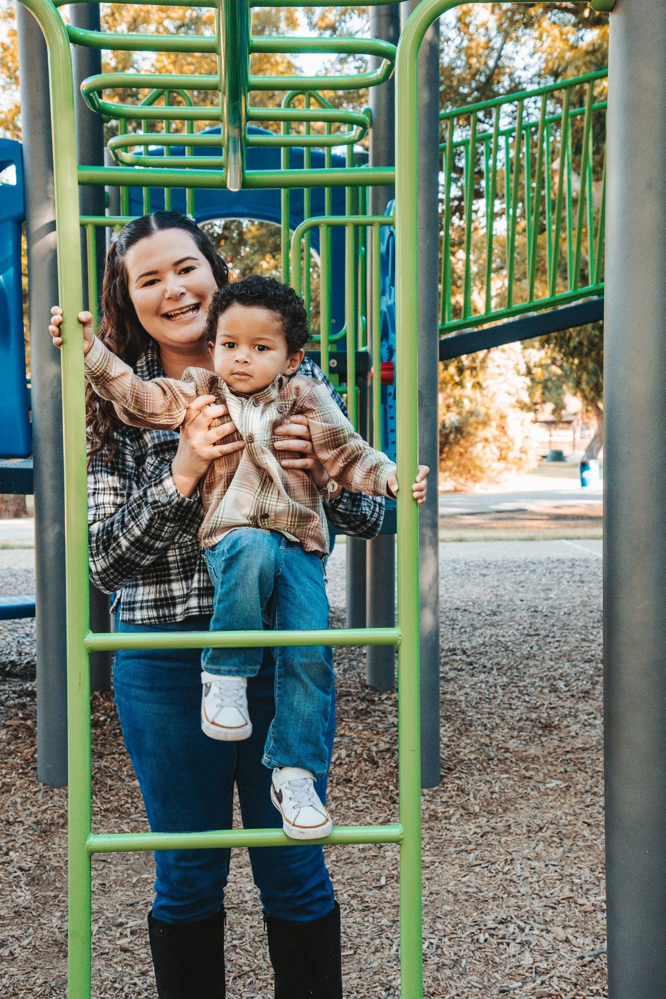 A woman with long brown hair smiling and holding a young boy with curly hair on a green playground ladder, with trees and a park in the background.