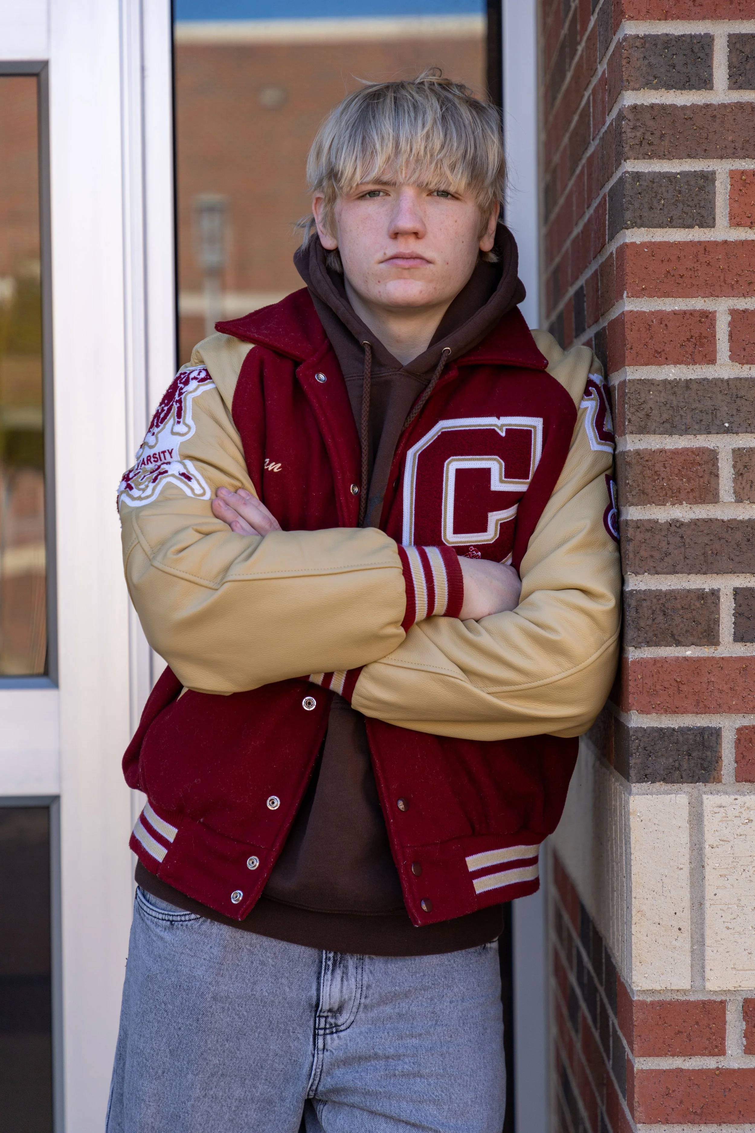 A teenage boy with blond hair and a serious expression, wearing a varsity jacket with a large letter C, standing with arms crossed against a brick wall.