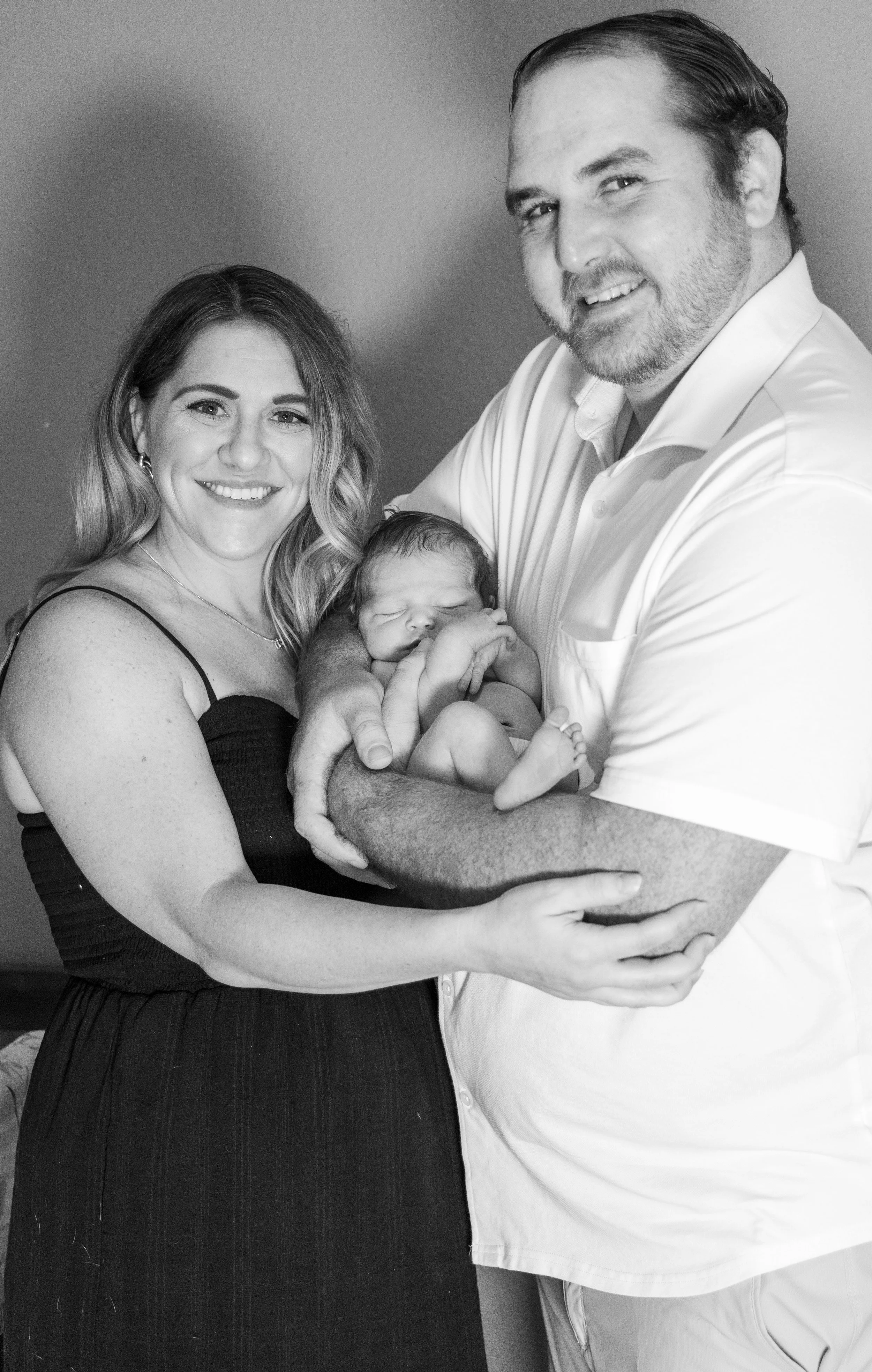 A black and white photo of four people: a smiling woman, a man holding a sleeping baby, and a person with only part of their body visible, all standing close together against a plain background.