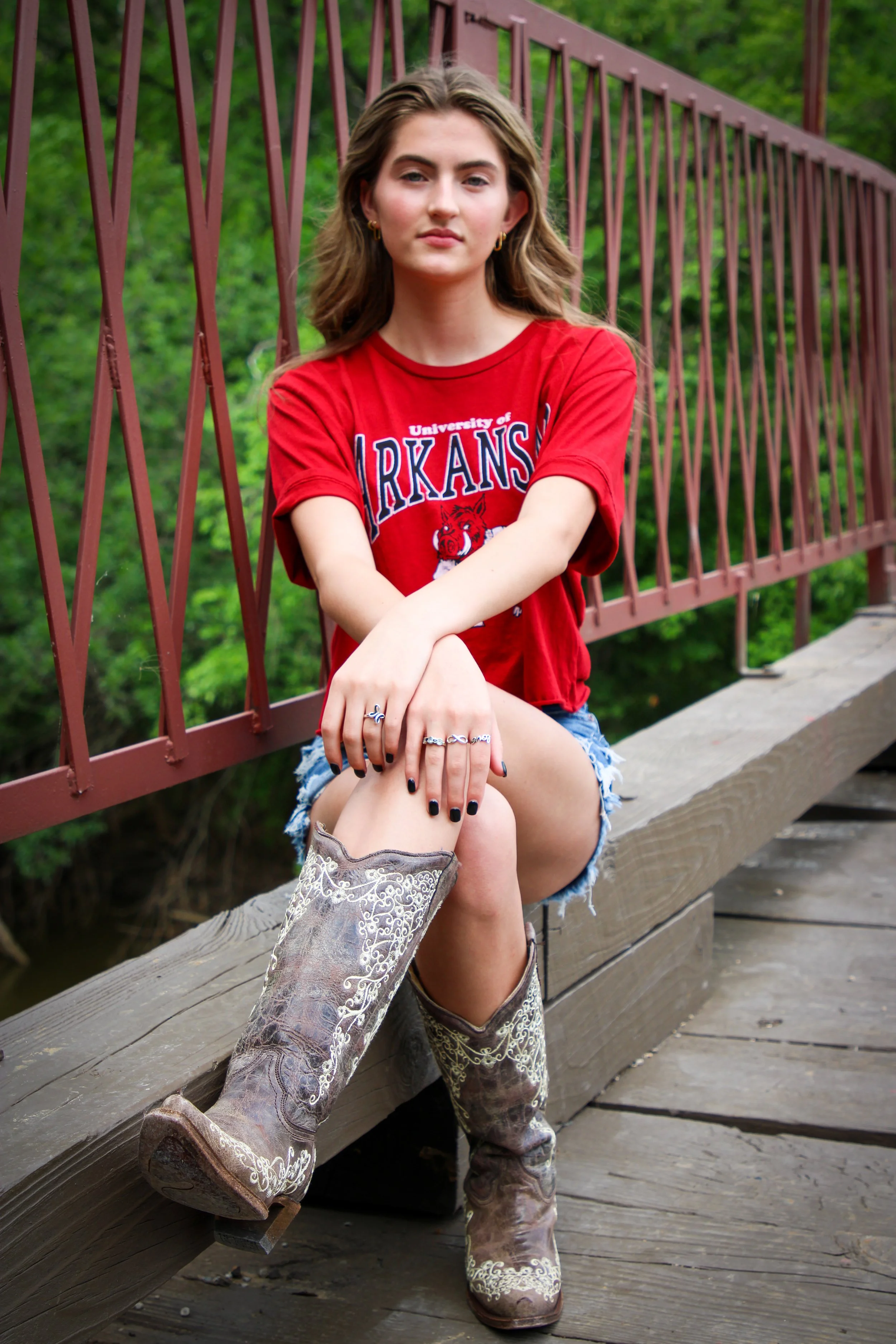 Young woman sitting on a wooden bridge with a red railing, wearing a red University of Arkansas T-shirt, distressed denim shorts, and cowboy boots, with green trees in the background.