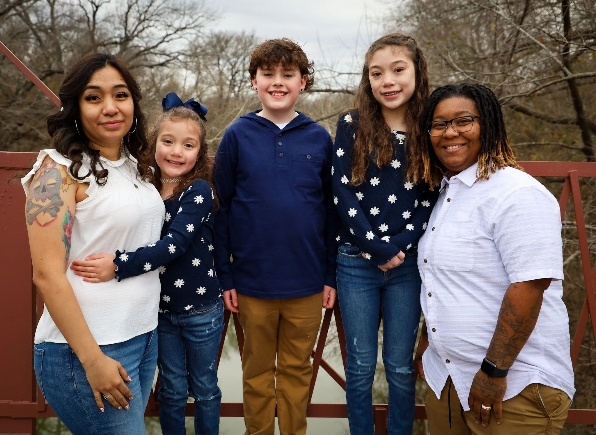 Six people standing on a bridge outdoors, smiling at the camera, with trees and a body of water in the background.