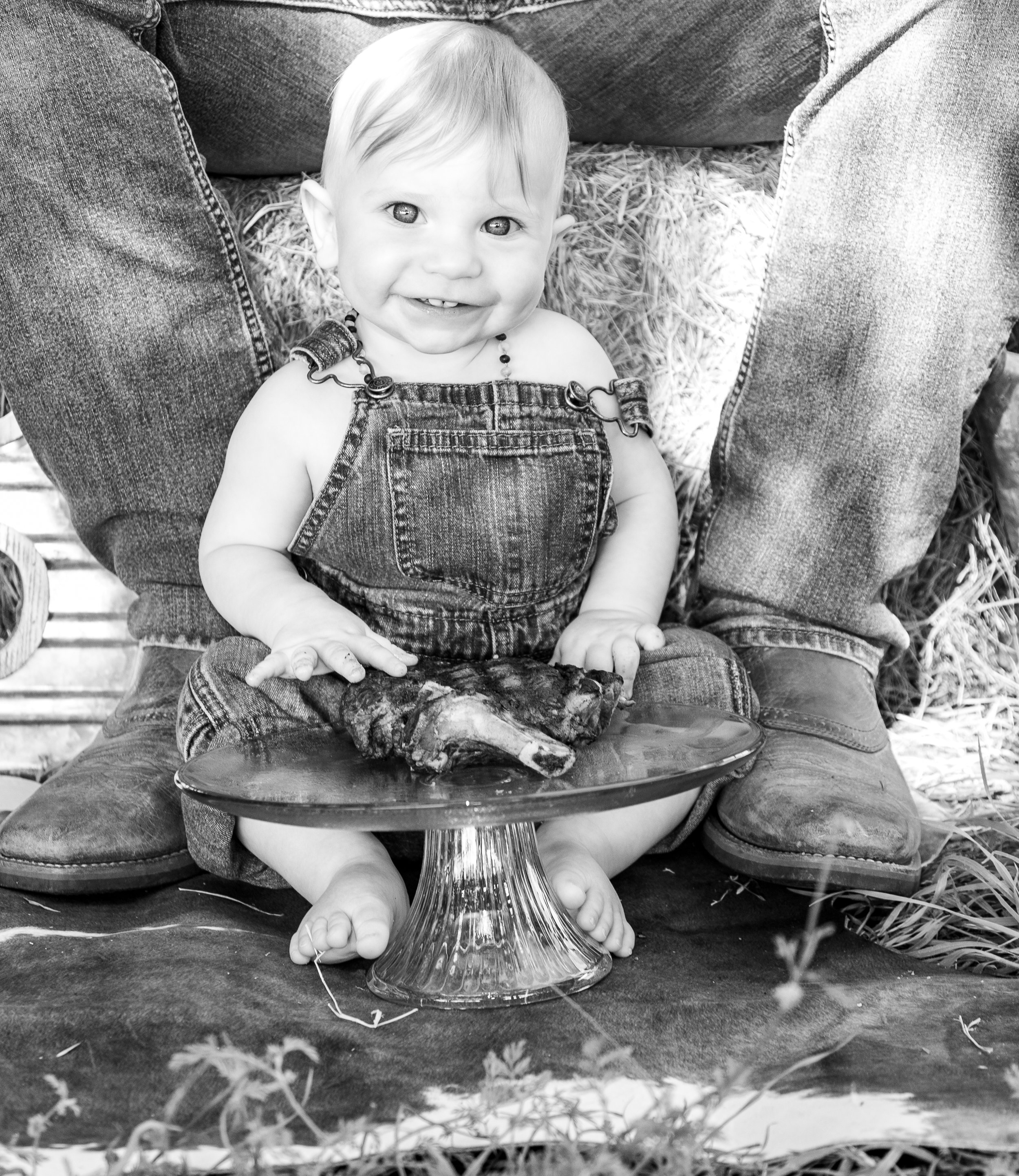 A young child sitting cross-legged on the ground, smiling, with a piece of meat on a glass cake stand in front of them. The child is wearing denim overalls and is surrounded by an adult wearing jeans and boots.