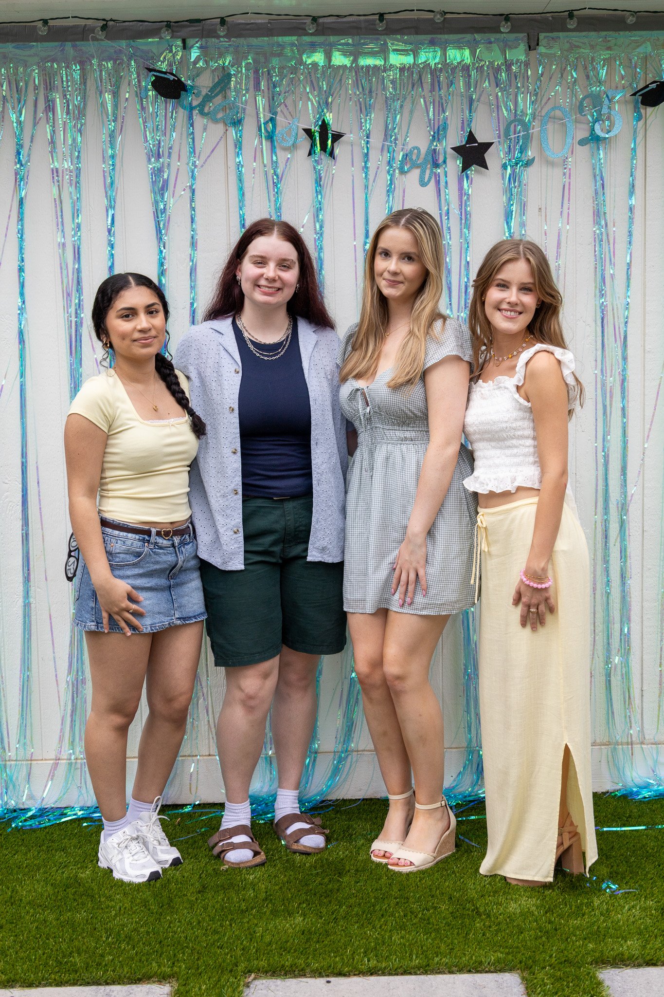 Four young women standing together at a celebration with iridescent decorations and a 'Congratulations' banner in the background.