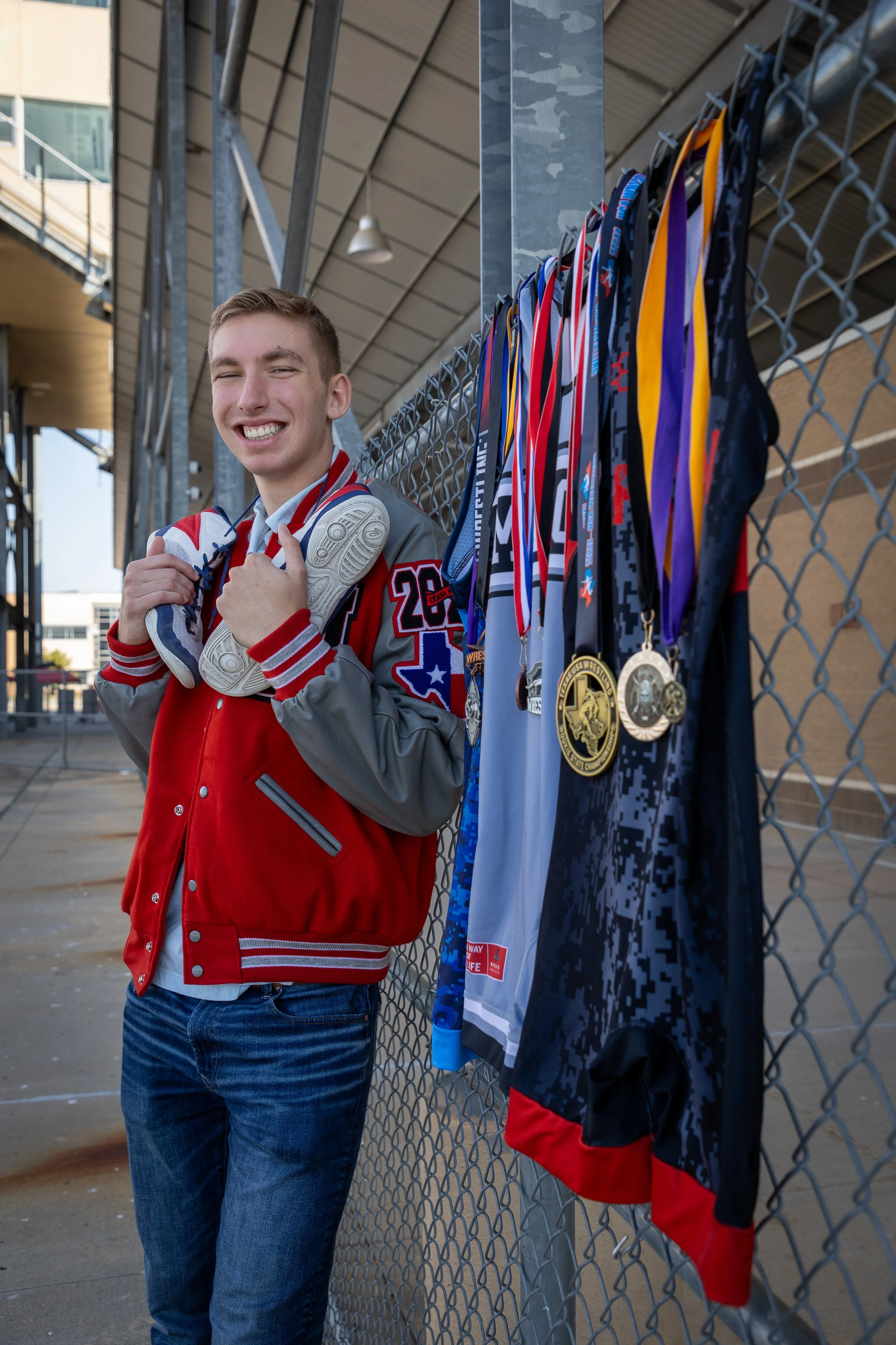 A young man in a red and gray sports jacket holding a pair of white sneakers stands next to a row of medals and athletic uniforms hanging on a fence, outside a sports facility.