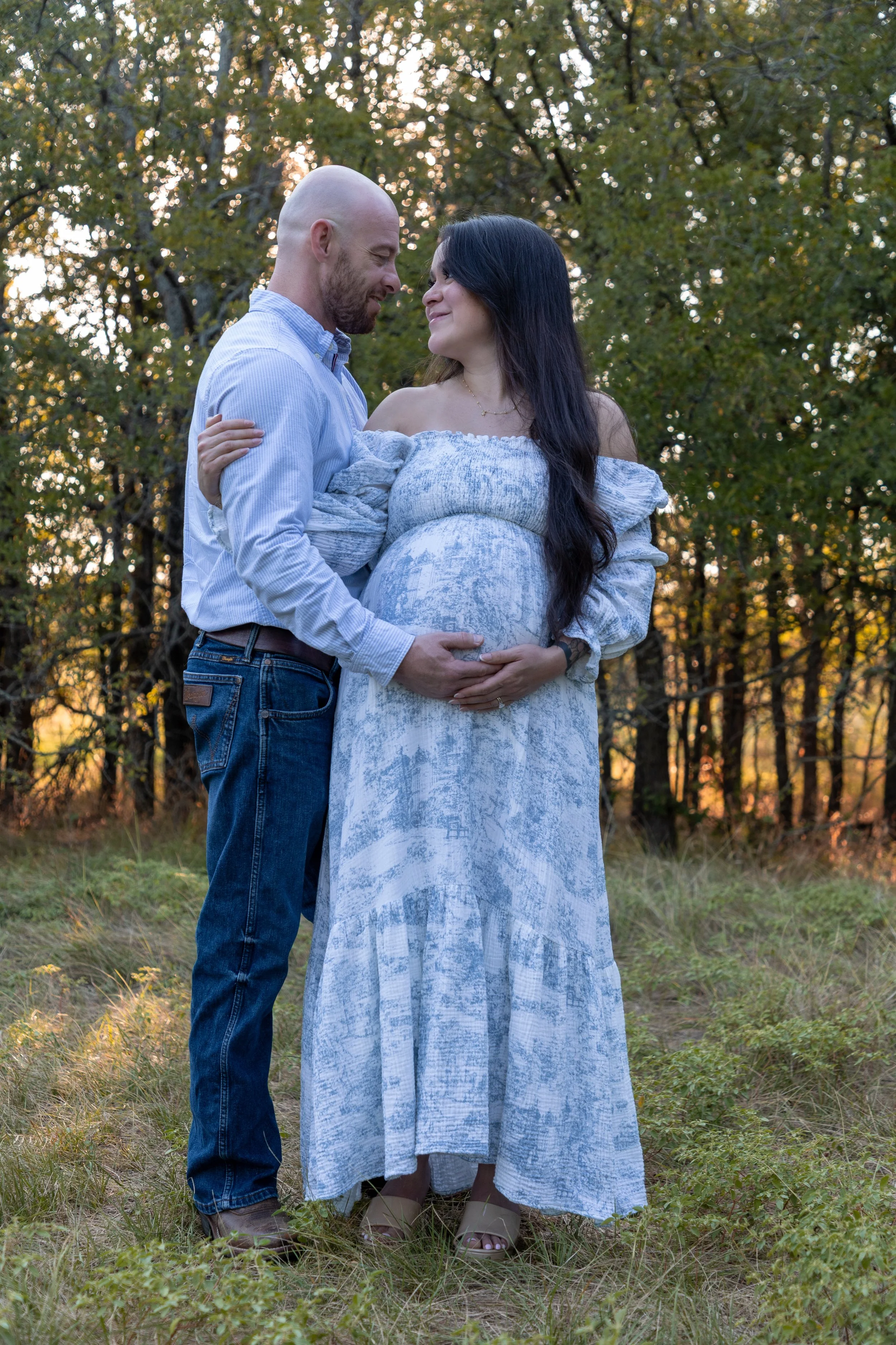 A pregnant woman in a long blue and white patterned dress and a man in a light blue shirt and jeans standing close in a grassy outdoor setting during sunset, affectionately touching her belly.
