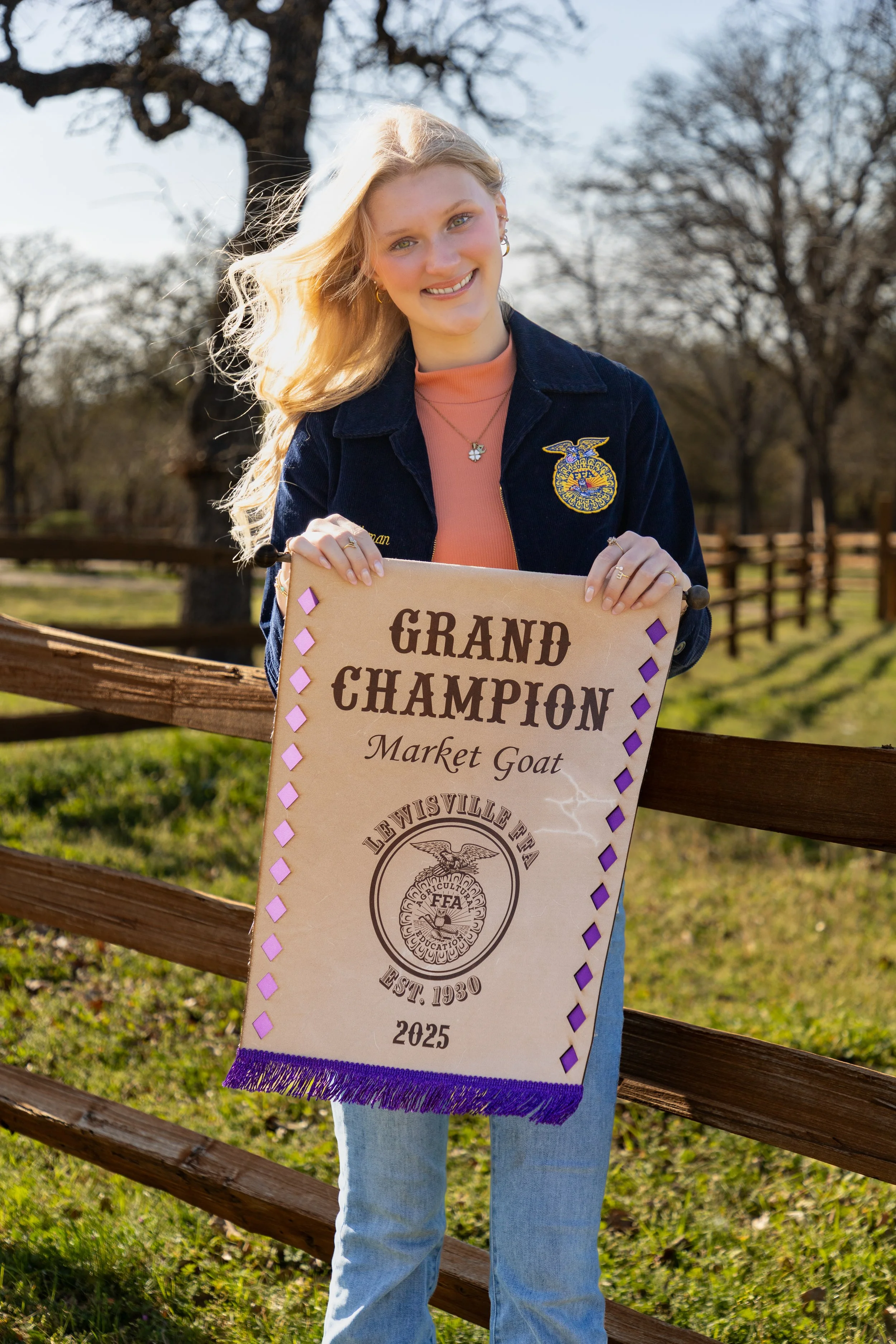 A young woman with long blonde hair smiling outdoors, holding a banner that reads 'Grand Champion Market Goat' at a livestock or agricultural fair, with trees and a wooden fence in the background.