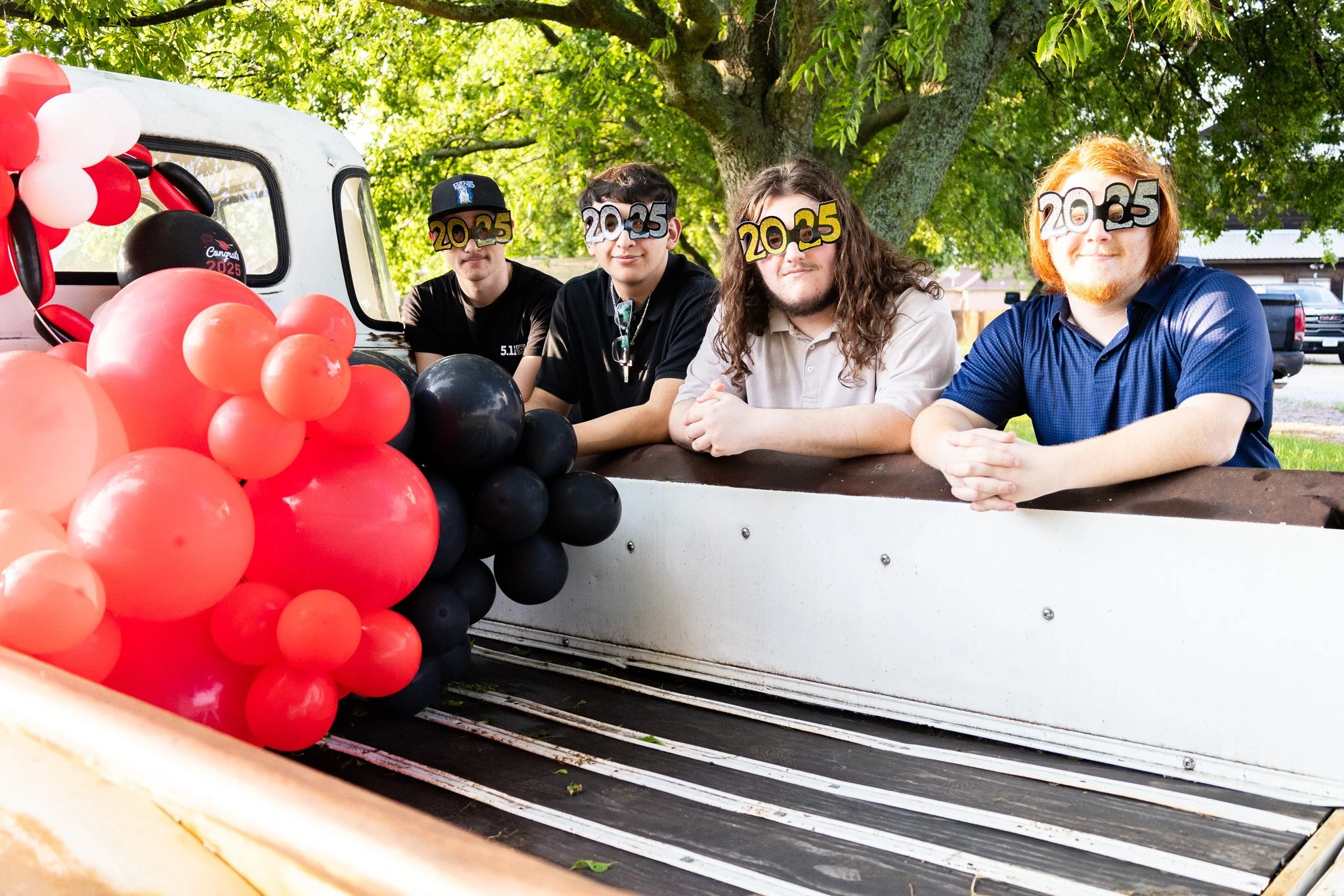 Four young men in casual clothing sitting inside a vintage pickup truck decorated with black, red, and white balloons, with the numbers "20-25" on their faces, under a large leafy tree.