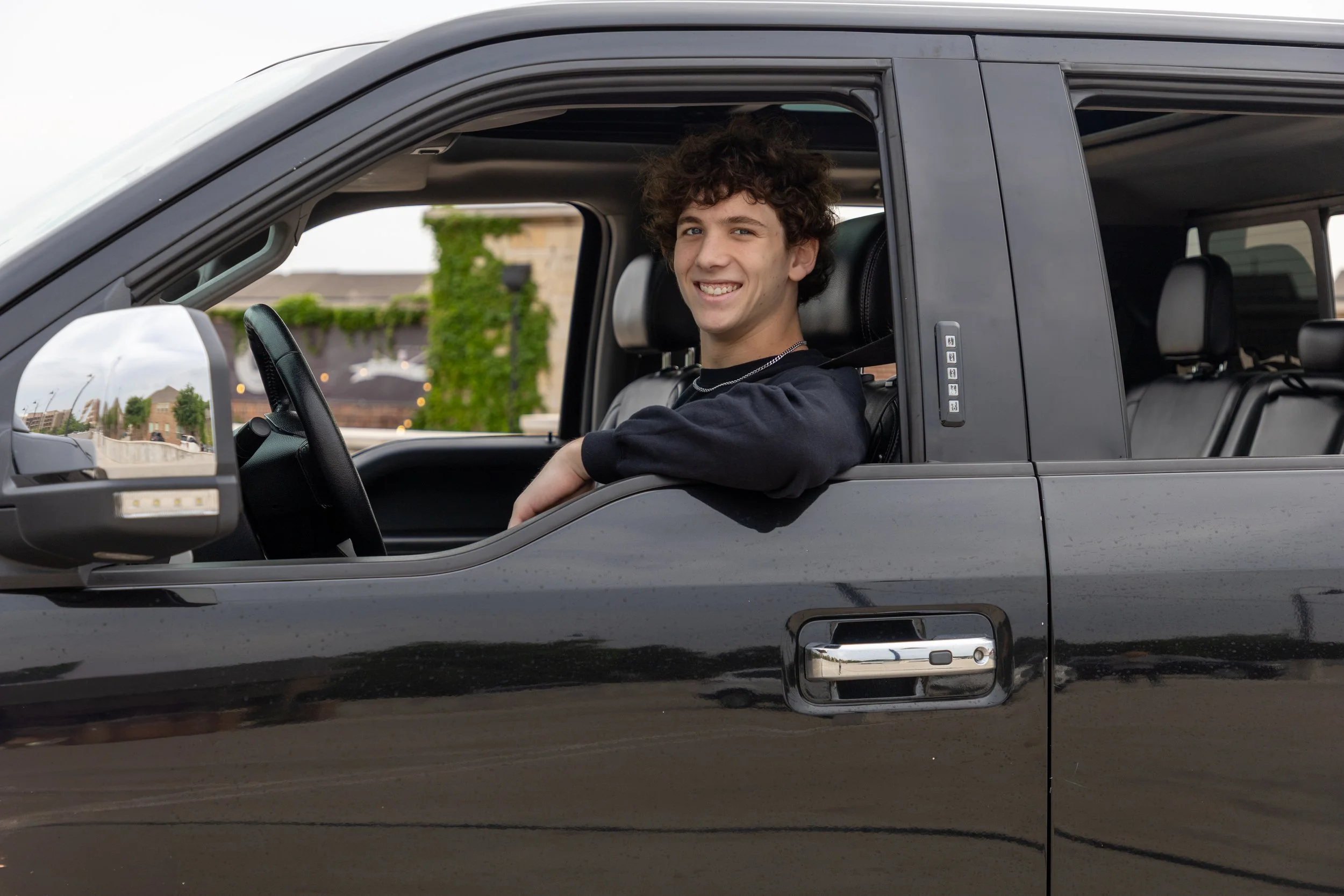 A young man with curly hair smiling while sitting in the driver's seat of a black pickup truck on a cloudy day.