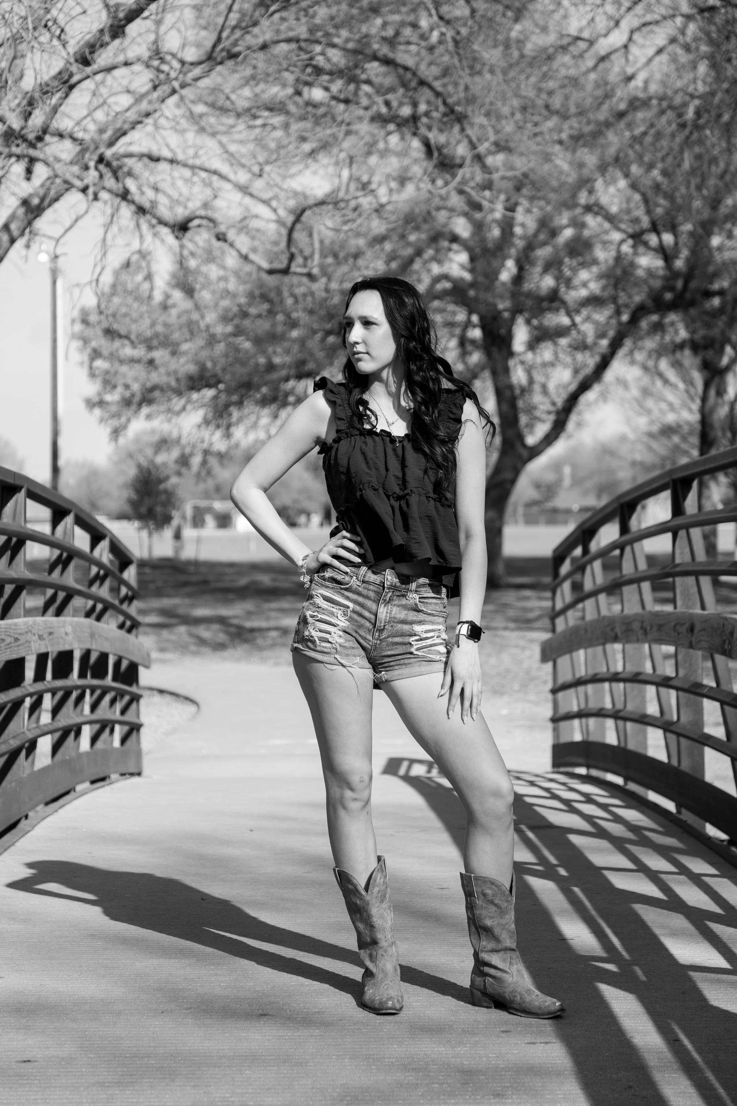 A woman standing on a bridge outdoors, wearing a sleeveless top, ripped shorts, and cowboy boots, with her left hand on her hip and looking to the left, in a black-and-white photo.