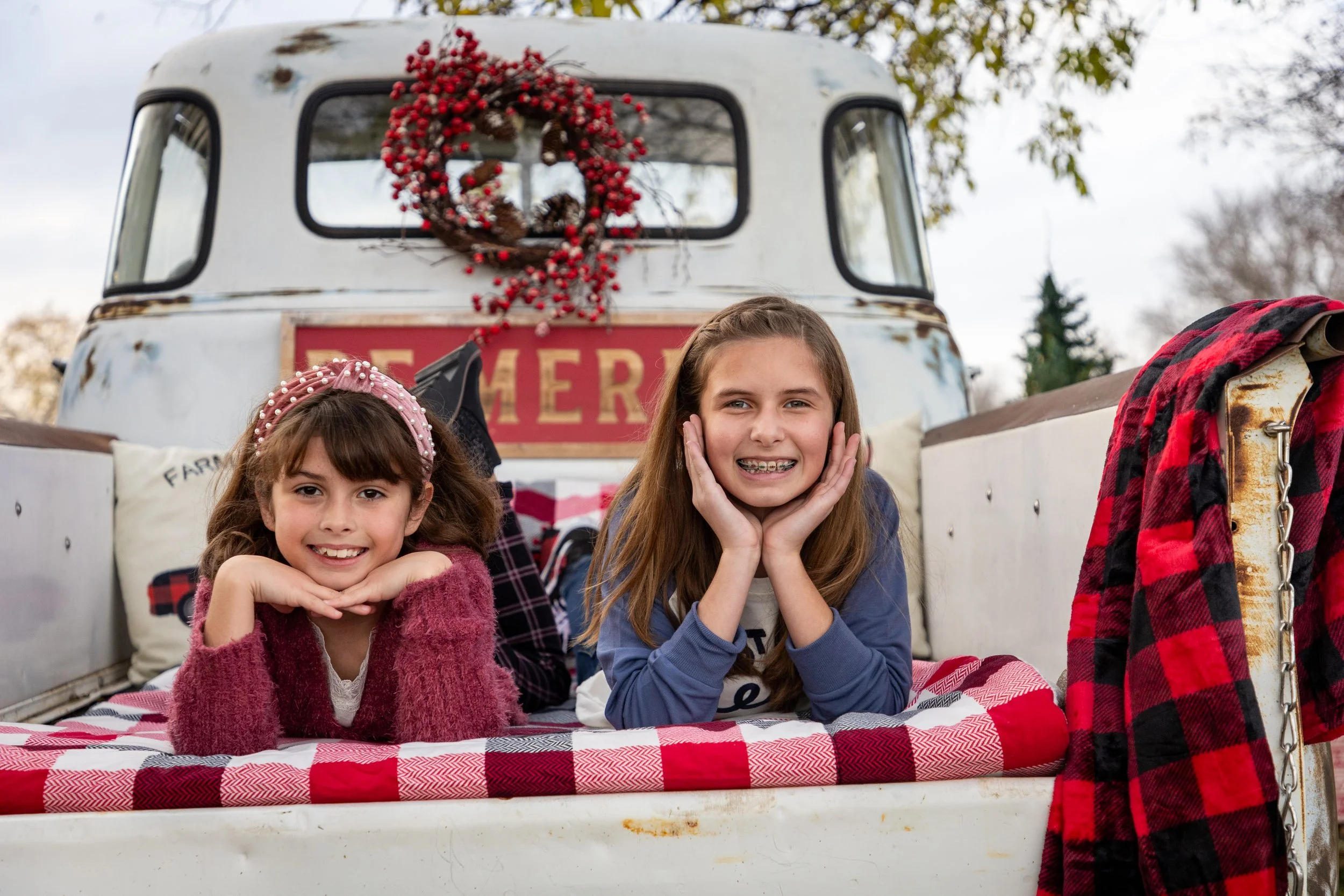 Two girls lying on a red and white checkered blanket in the back of an old white truck decorated with a Christmas wreath, with trees and cloudy sky in the background.