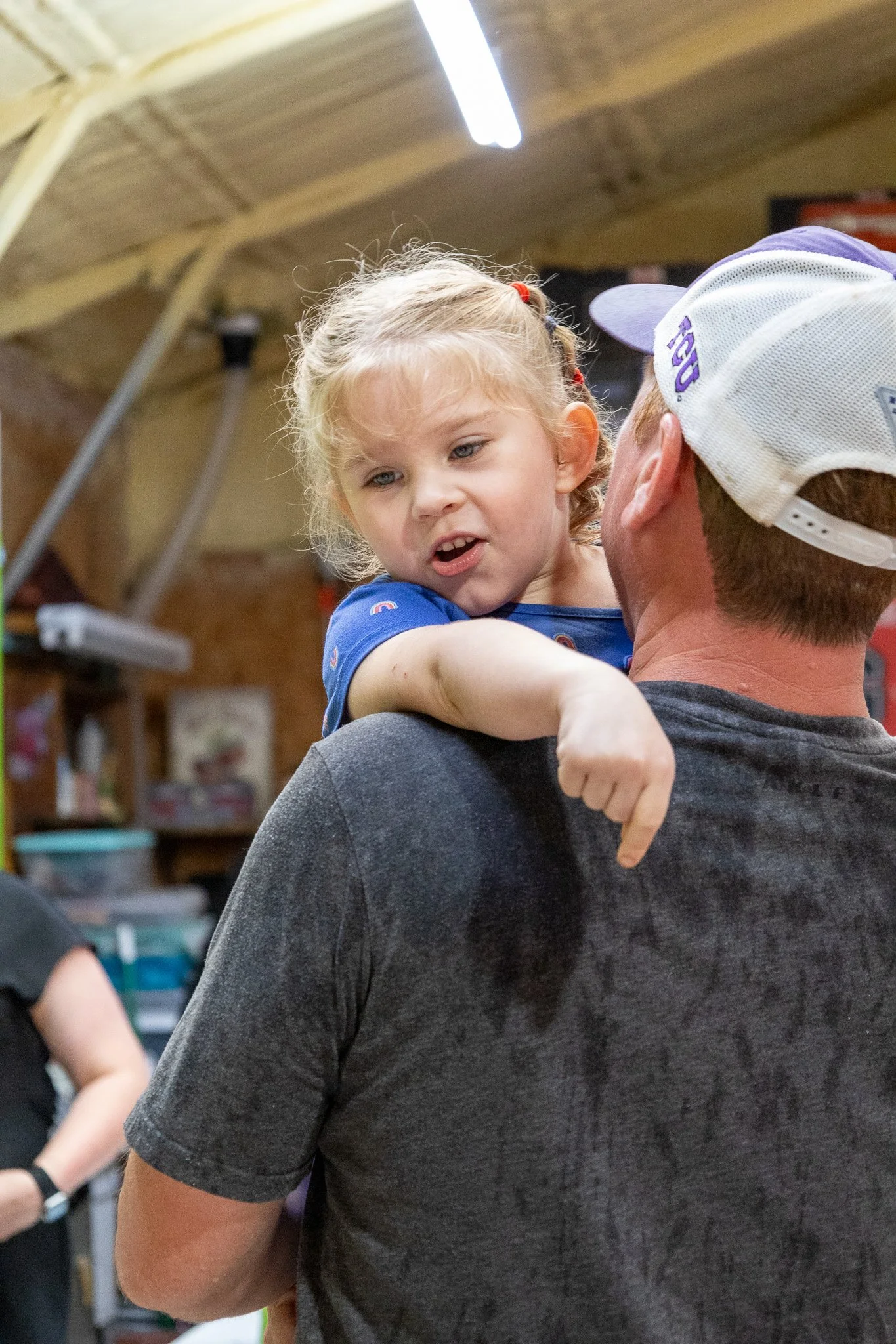 A young girl with curly blonde hair, wearing a blue shirt, being carried on a man's shoulder in an indoor setting, possibly a garage or store, with shelves and objects in the background.