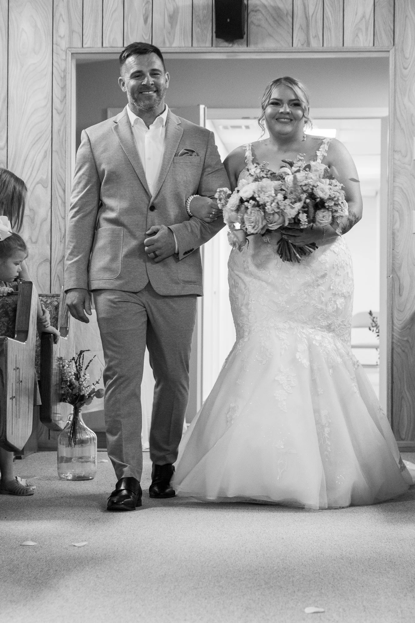 Black and white photo of a bride holding a bouquet of flowers walking down the aisle with a man, likely her father, in a suit, inside a church with wooden walls.