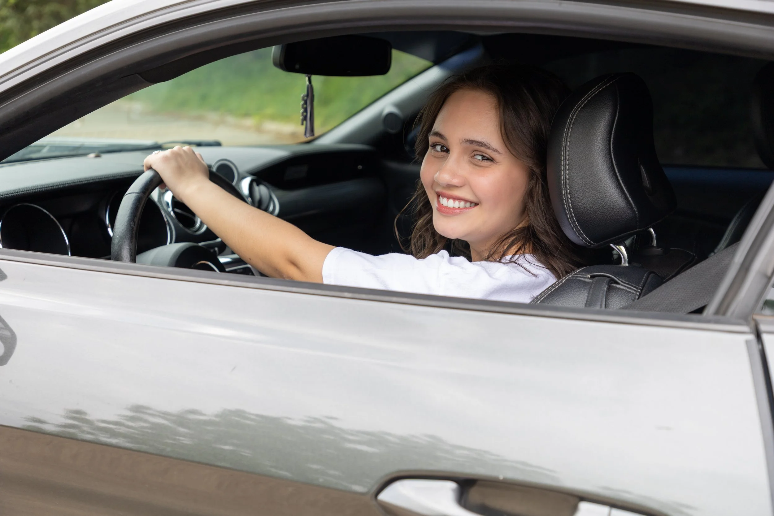 A young woman with brown hair and a white shirt sitting in the driver's seat of a silver car, smiling and looking at the camera, with her left hand on the steering wheel.