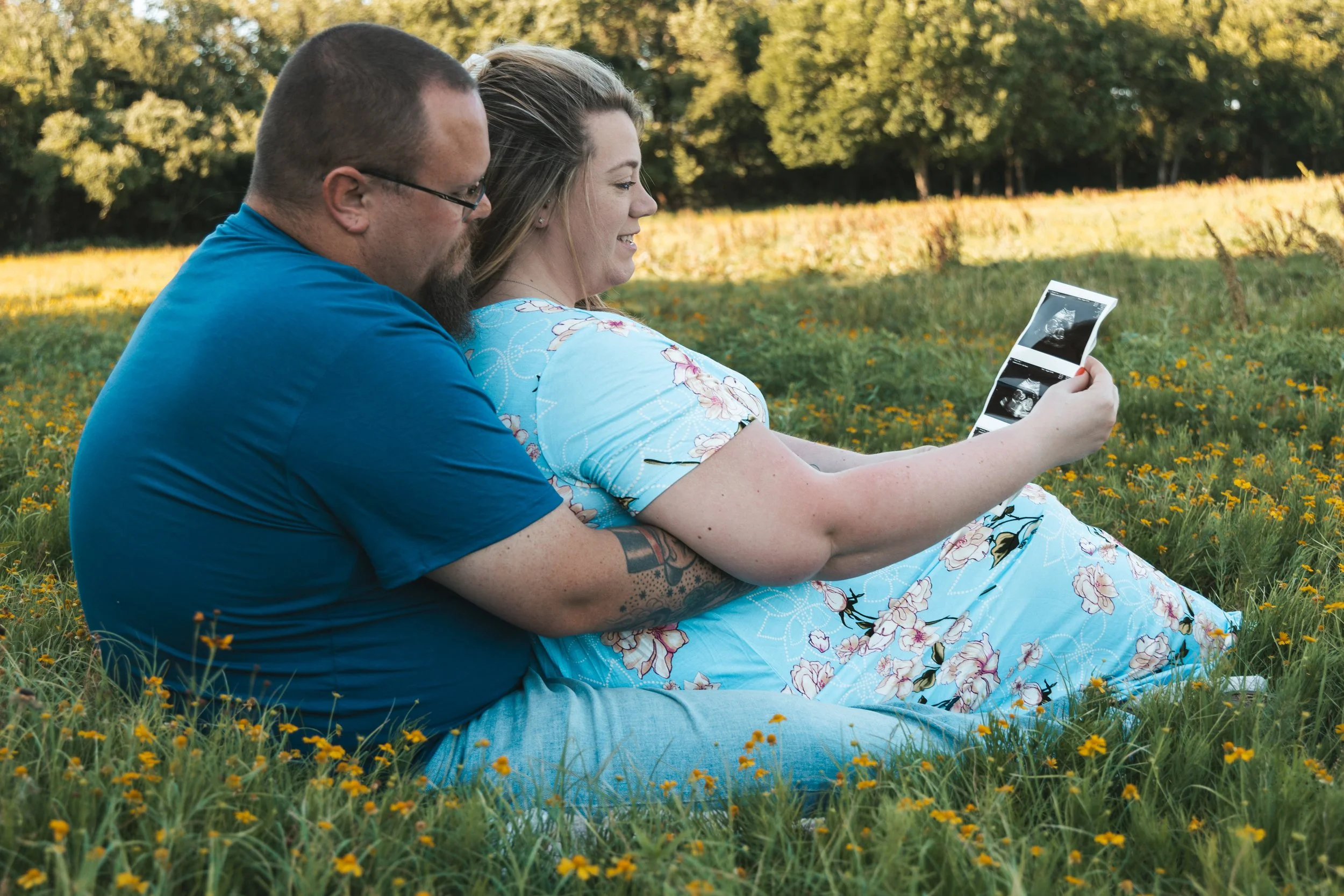 A couple sitting in a field of yellow flowers, looking at ultrasound images, with trees and sunlight in the background.