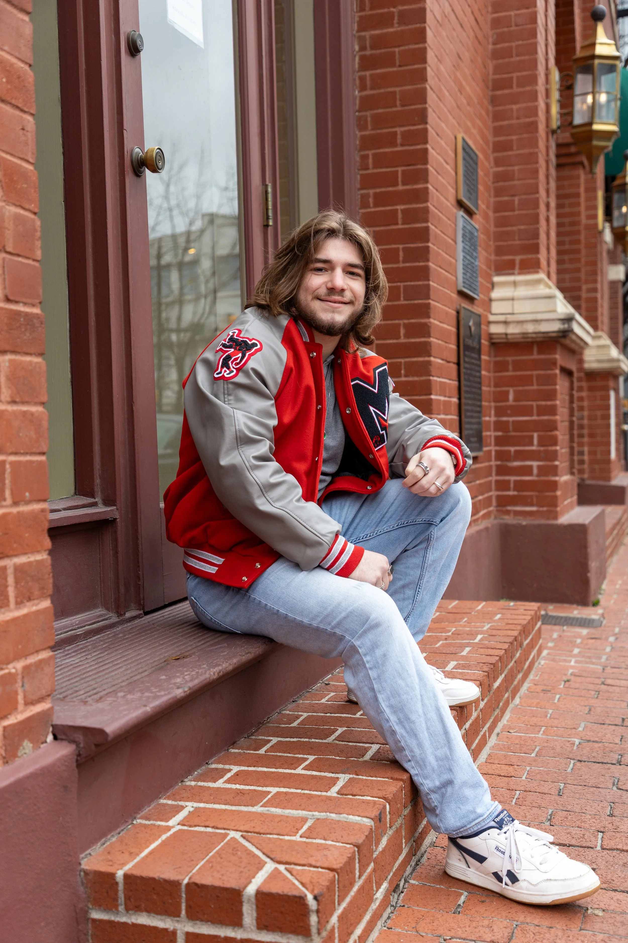 A young man with shoulder-length hair and a beard sitting on a brick ledge outside a red brick building, smiling, wearing a red and gray letterman jacket, light blue jeans, and white sneakers.