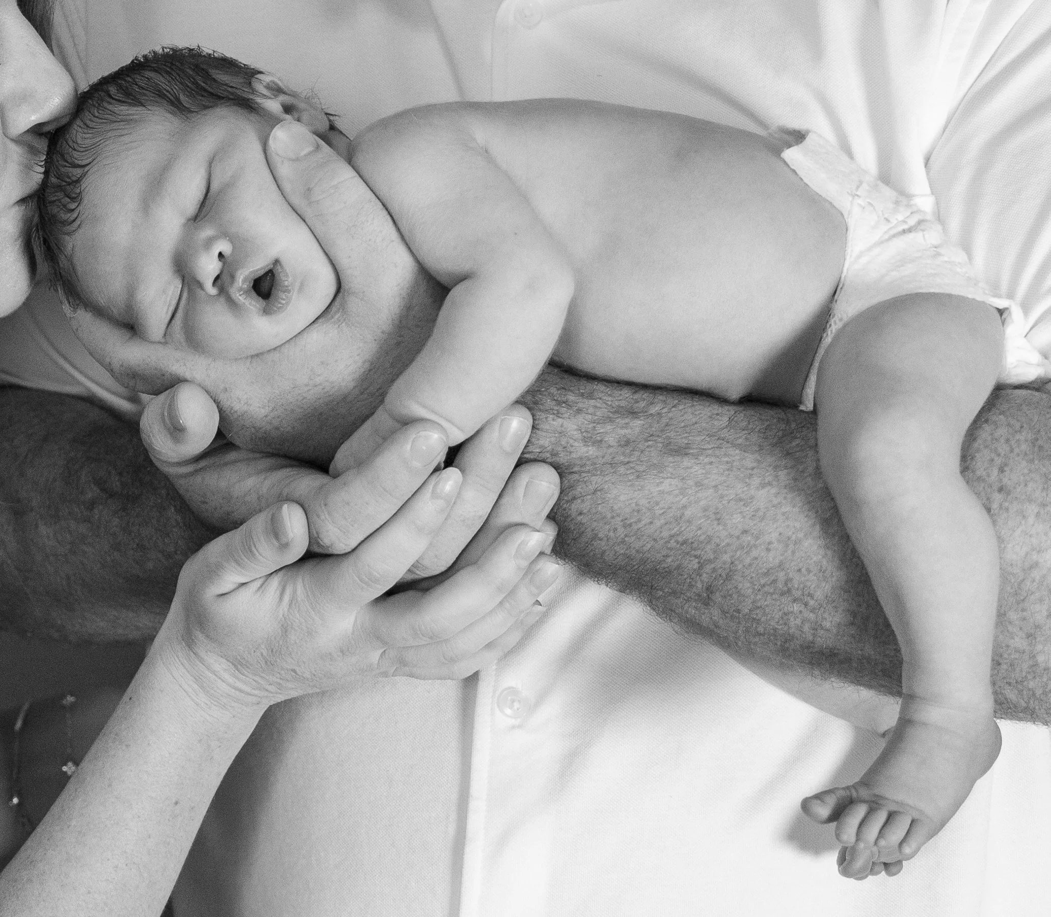 A newborn baby wearing a diaper, lying on a person's arm with adult hands holding the baby's head and arm, captured in black and white.