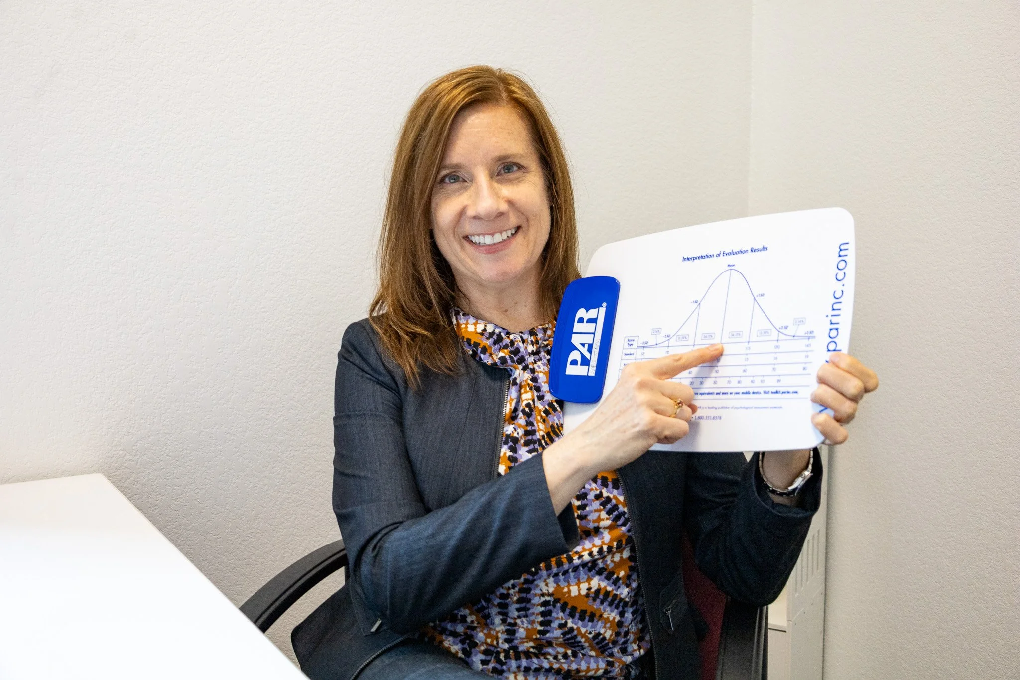 A woman with reddish-brown hair and a black blazer is holding a report with a chart, smiling at the camera.