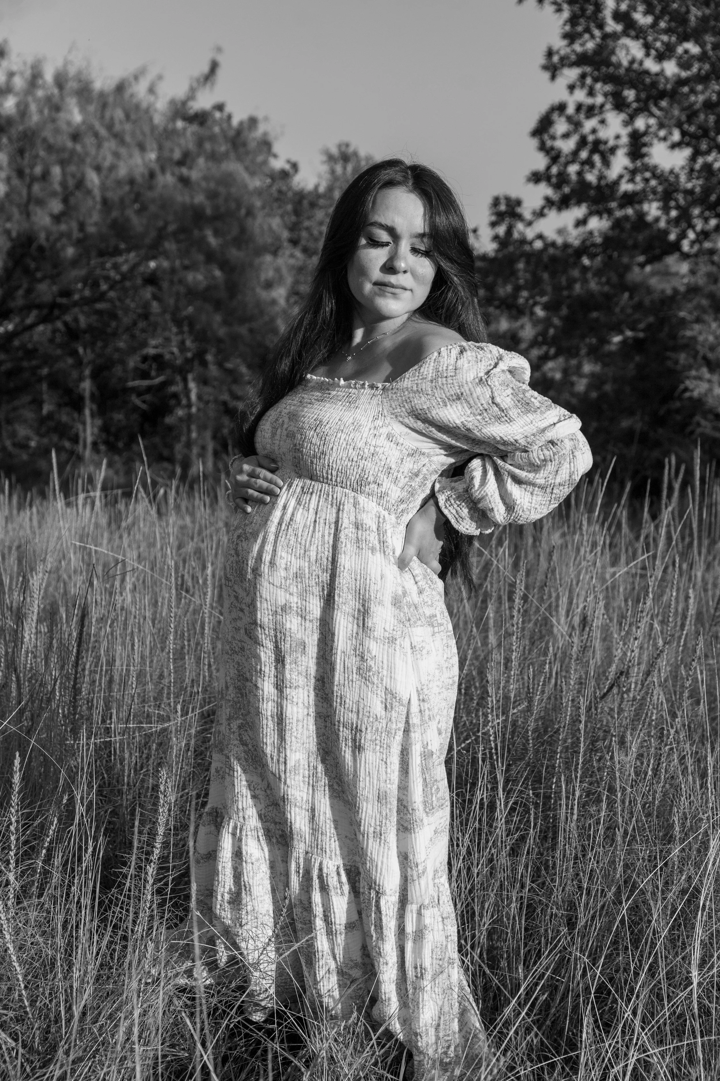 Black and white photograph of a young woman standing in a grassy field with trees in the background, wearing a patterned dress with puffed sleeves, looking down with a contemplative expression.