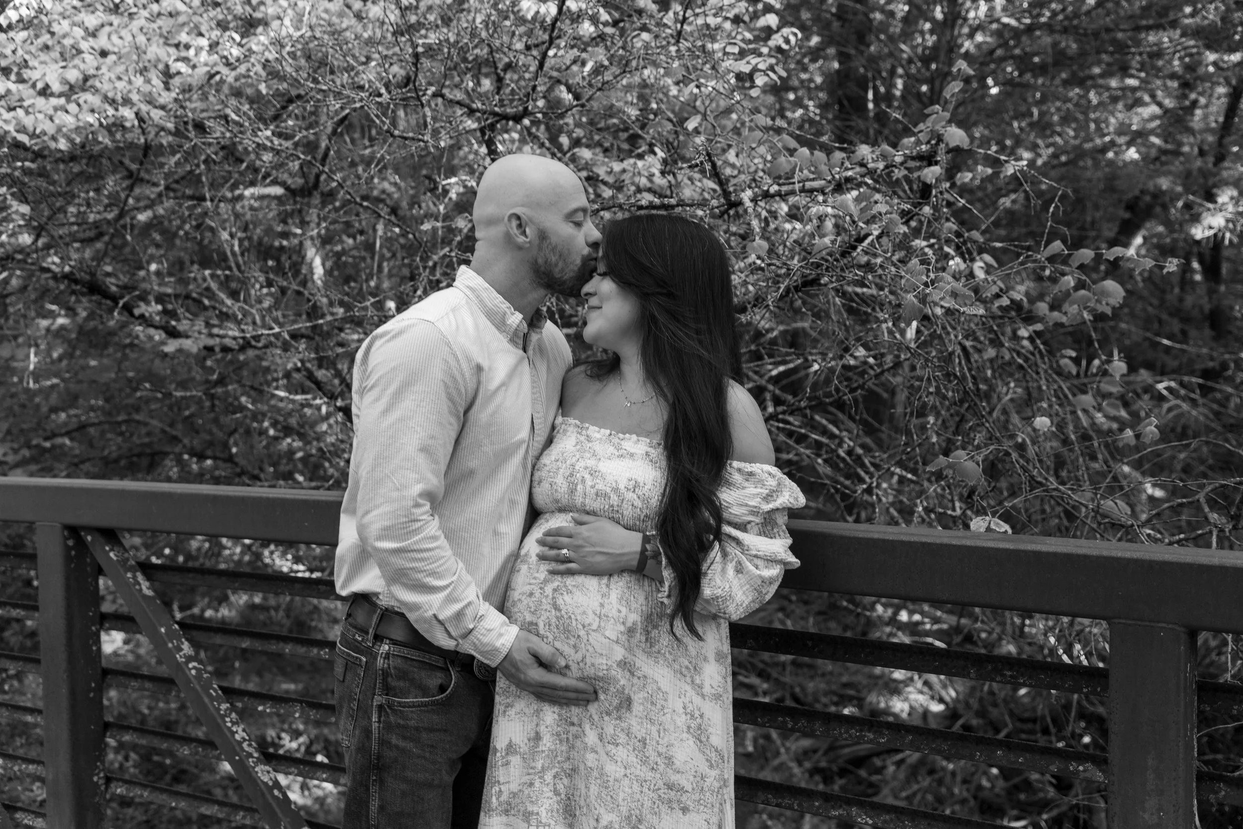 A black and white photo of a couple standing close on a bridge in a wooded area, with the man kissing the woman's forehead and the woman smiling with her hand on her belly.