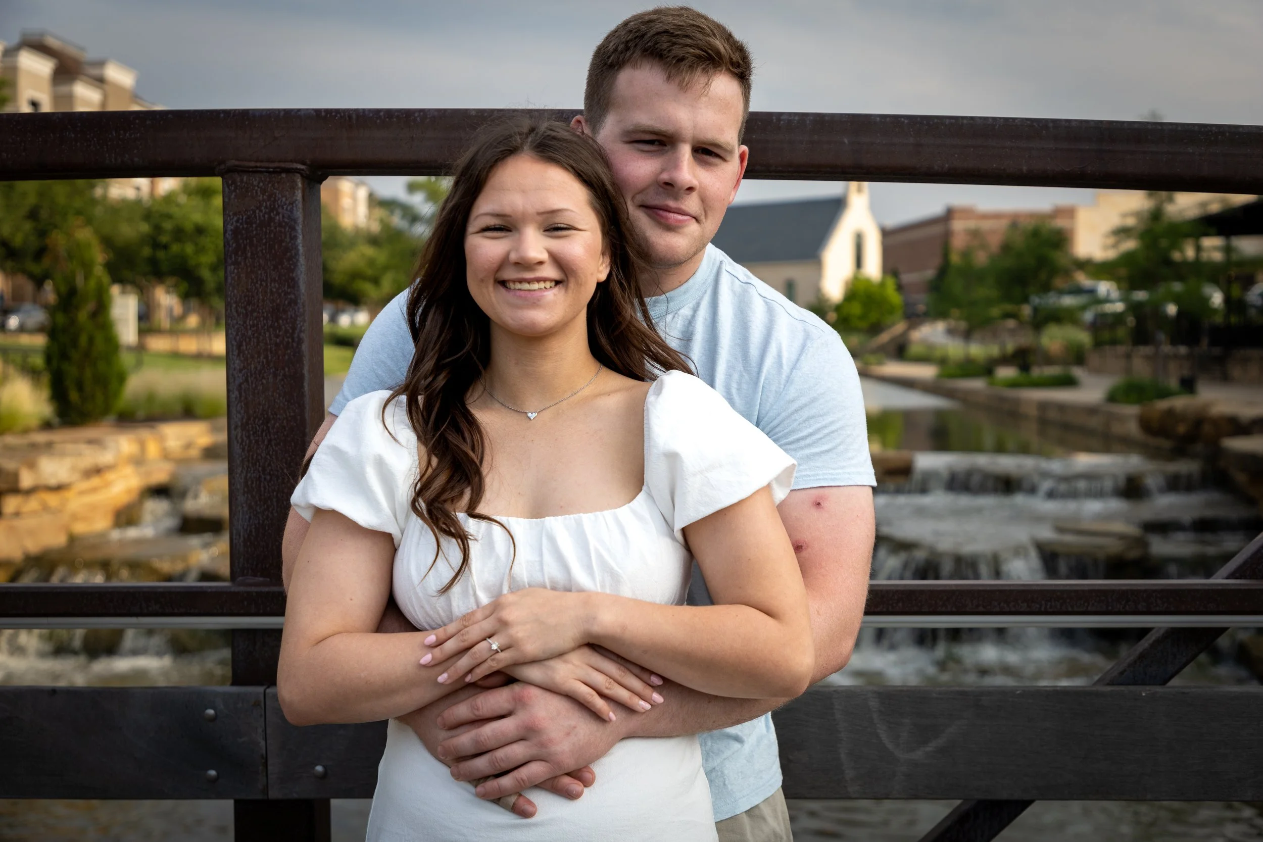 A smiling woman and a man standing behind her, on a bridge near a creek, with buildings and trees in the background.